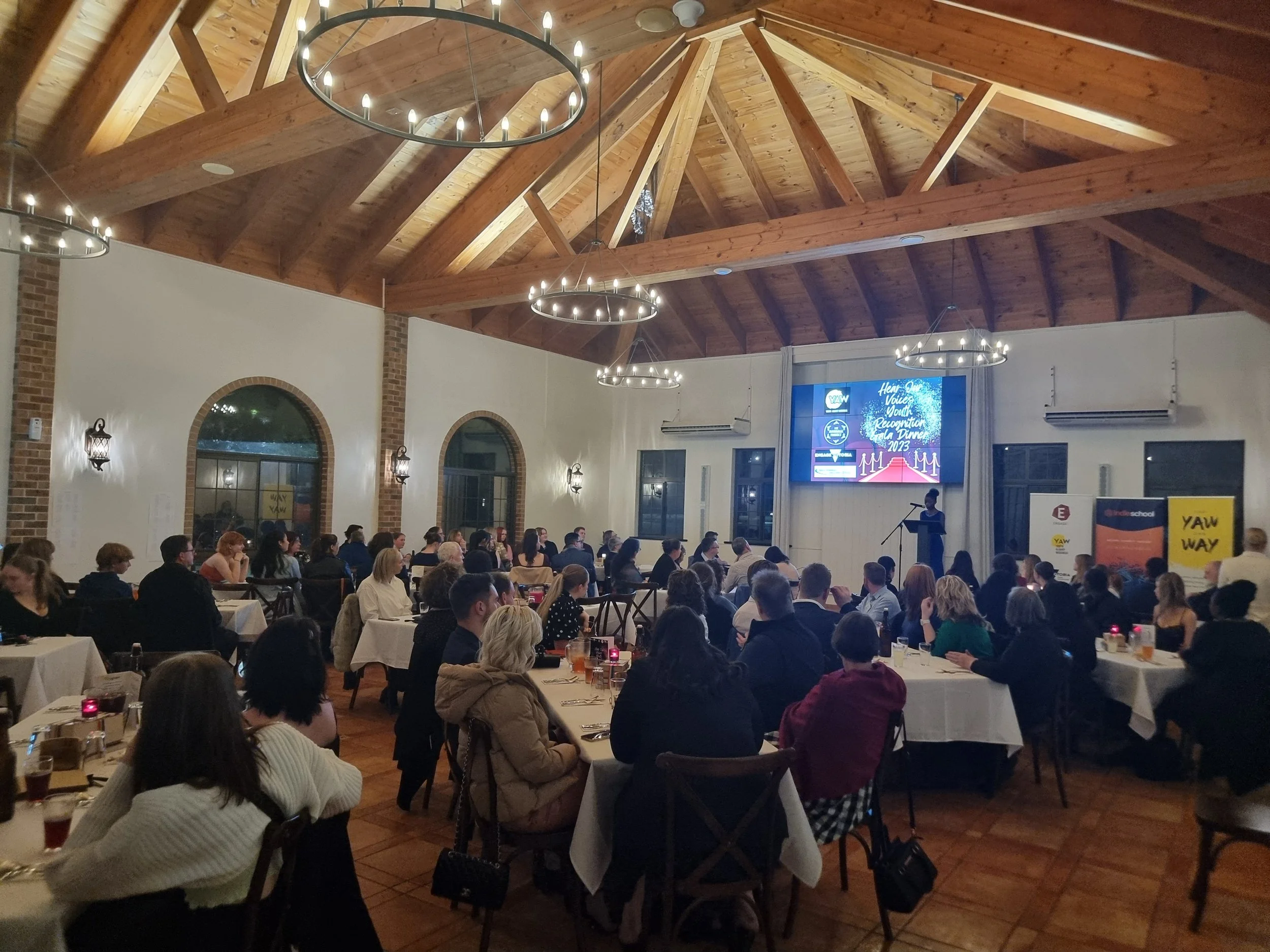 A large gathering of people seated at round tables during a formal event in a spacious room with a high wooden ceiling and chandeliers. A woman is speaking at a podium in front of a screen displaying a presentation.