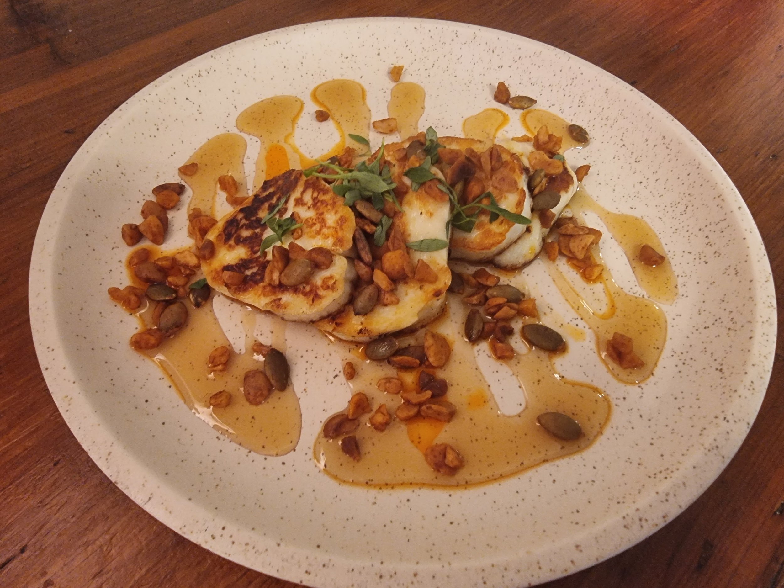 Grilled scallops topped with herbs, surrounded by toasted nuts and drizzled with honey on a white speckled plate, on a wooden table.