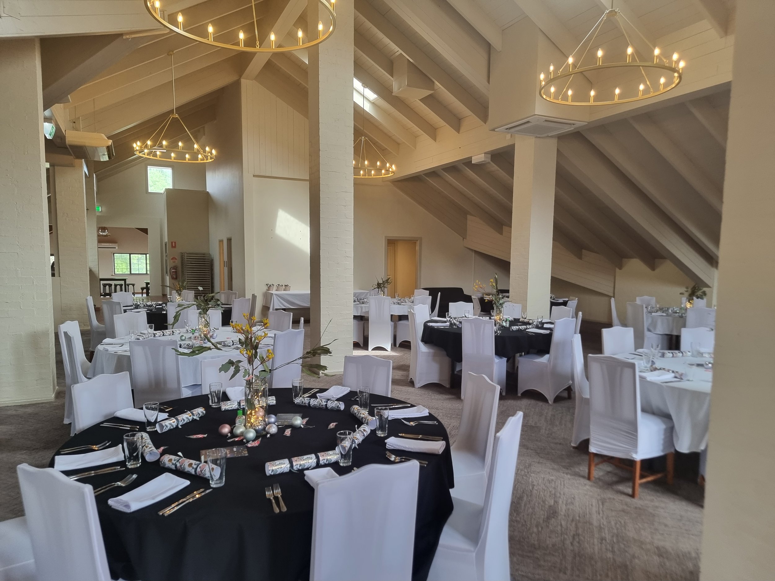 Event venue decorated with round tables covered in black and white tablecloths, floral centerpieces, and elegant place settings, under large chandeliers in a spacious room with vaulted ceilings.