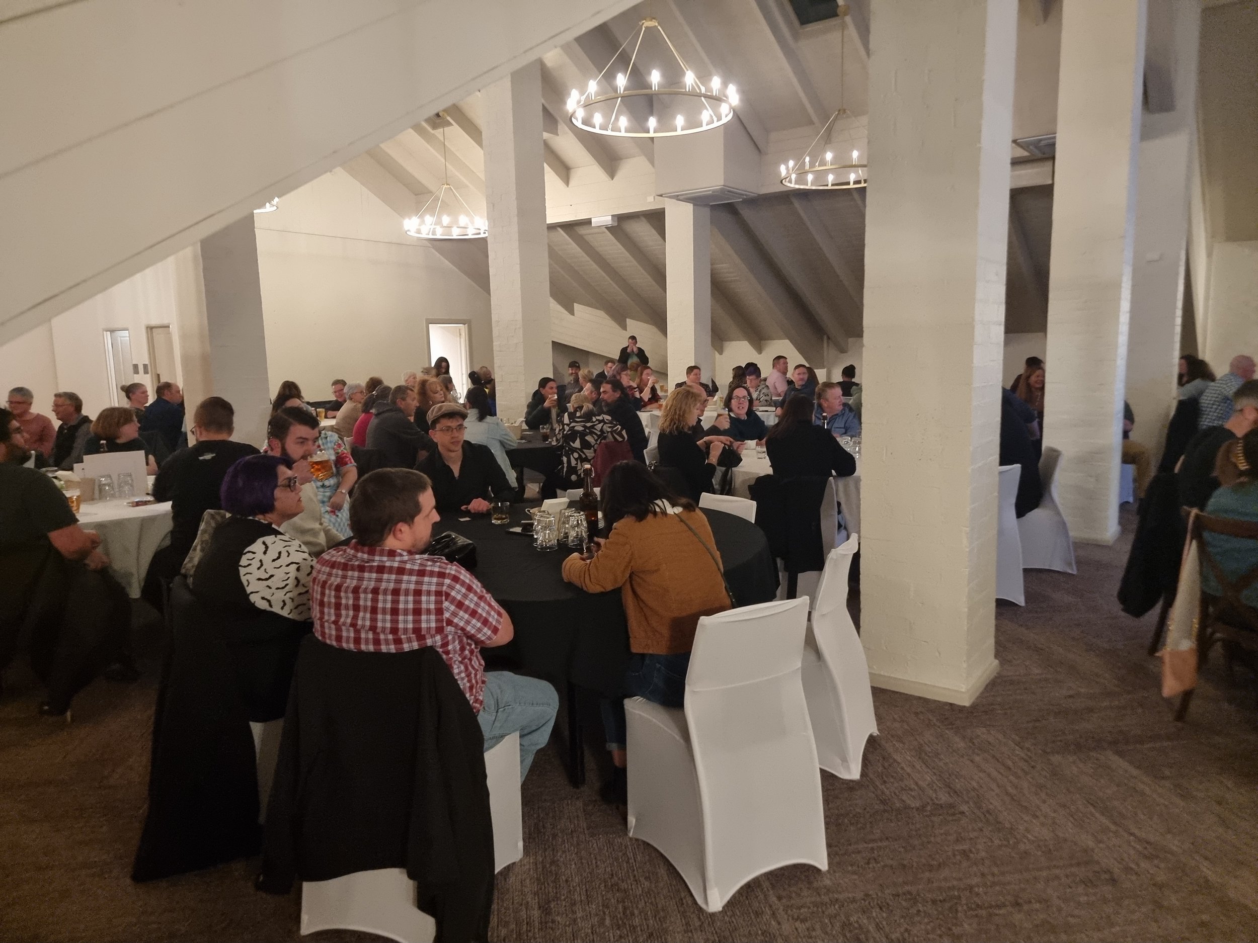 A large group of people gathering in a spacious banquet hall with white brick walls and high vaulted ceilings for a social event, seated at tables with black and white tablecloths.