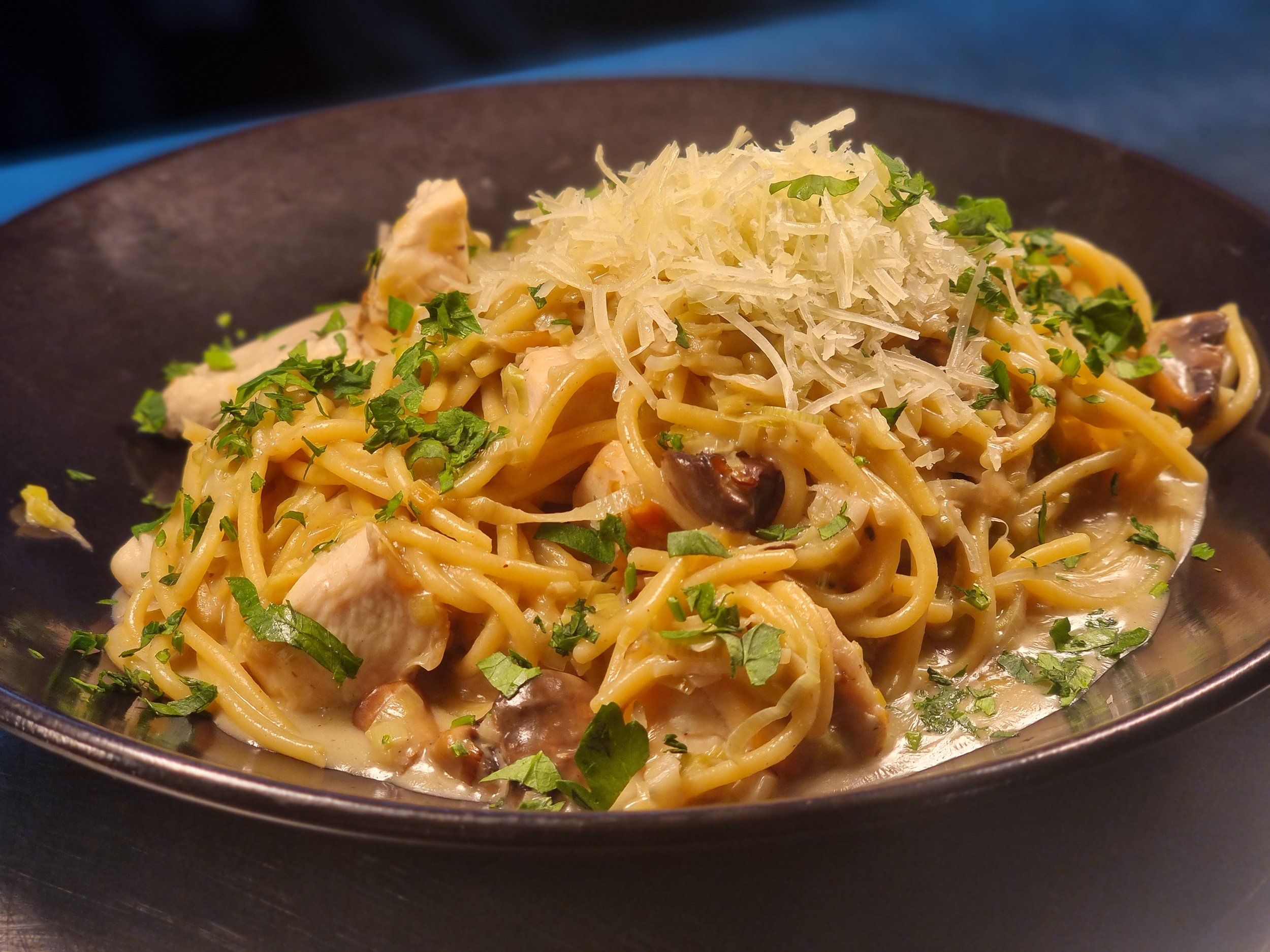 Spaghetti pasta with chicken, mushrooms, and grated cheese, garnished with chopped parsley in a black bowl.