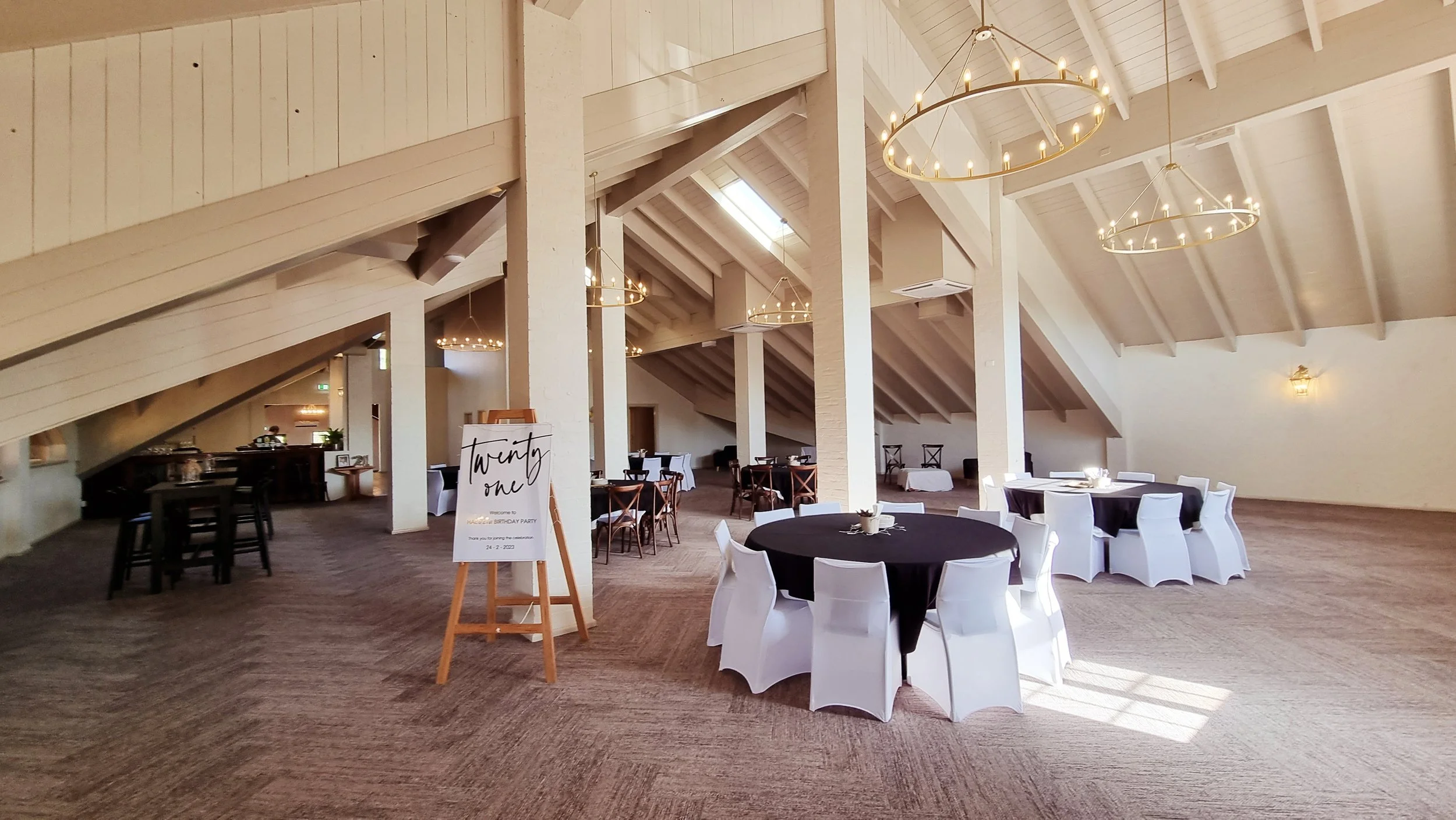 A spacious event hall with round tables covered in black or white tablecloths, chairs with white slipcovers, and large circular chandeliers hanging from a high vaulted ceiling with skylights. There is a sign on an easel indicating a birthday party ev