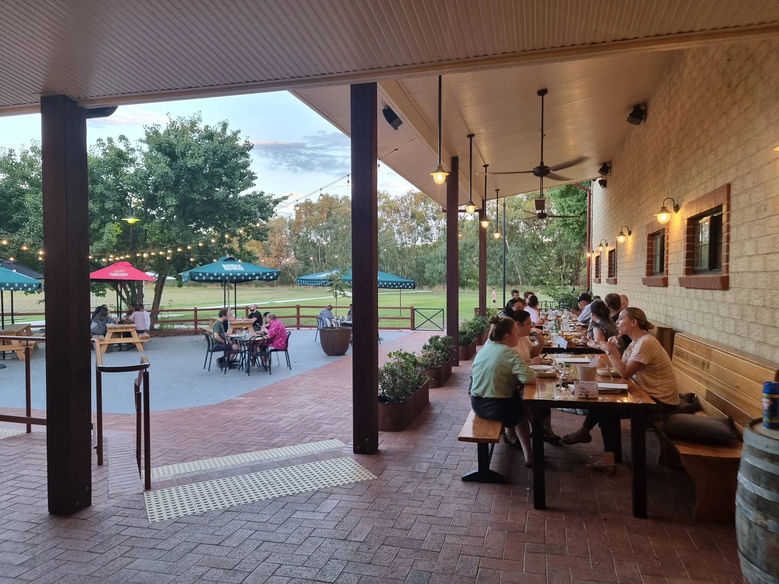 People dining inside and outside at a restaurant patio with string lights and umbrellas, surrounded by trees.