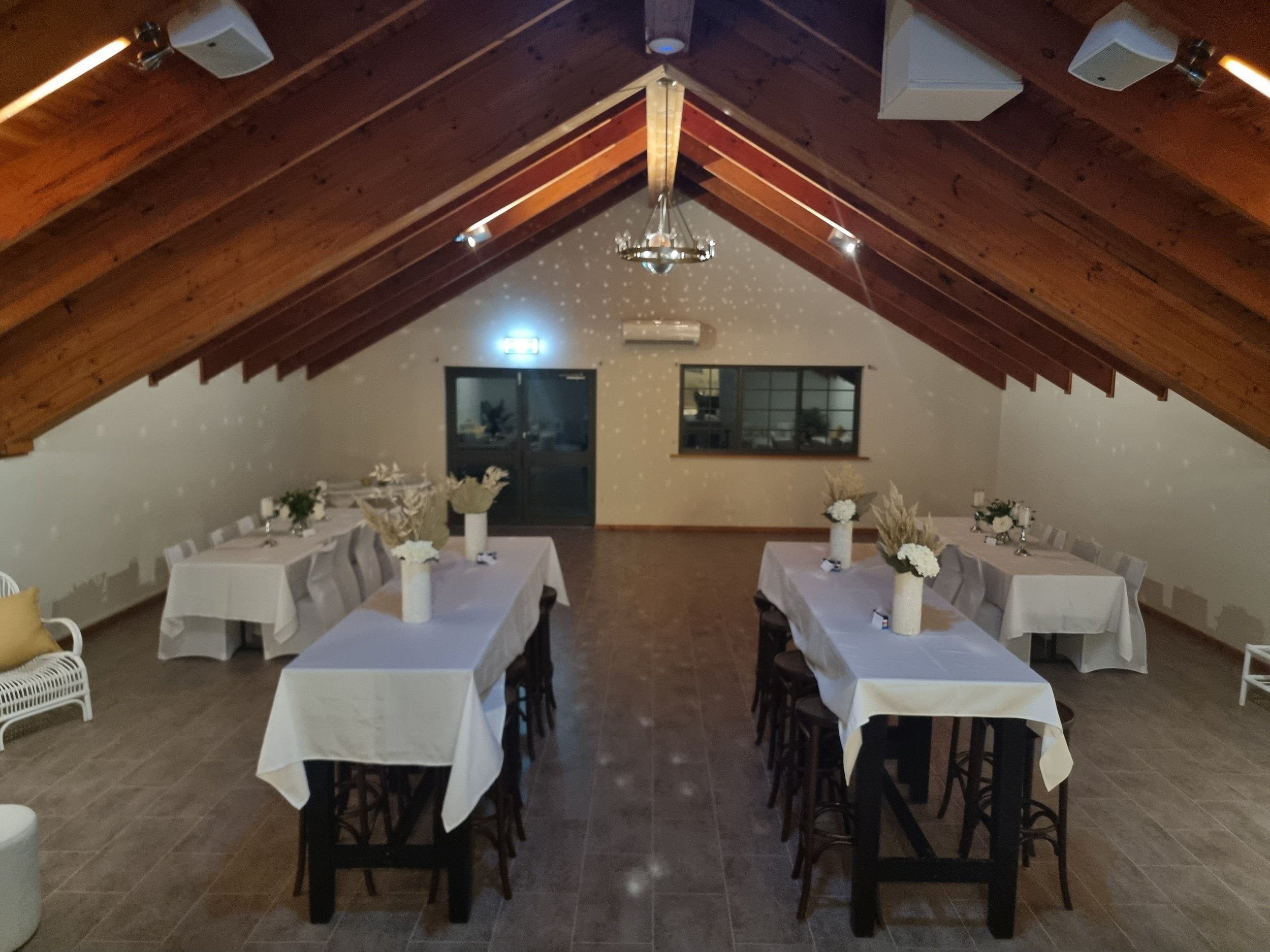 An indoor event space with a wooden vaulted ceiling, decorated for a formal gathering with white tablecloths, floral centerpieces, and chairs around rectangular tables, with soft lighting and star-like projections on the ceiling.