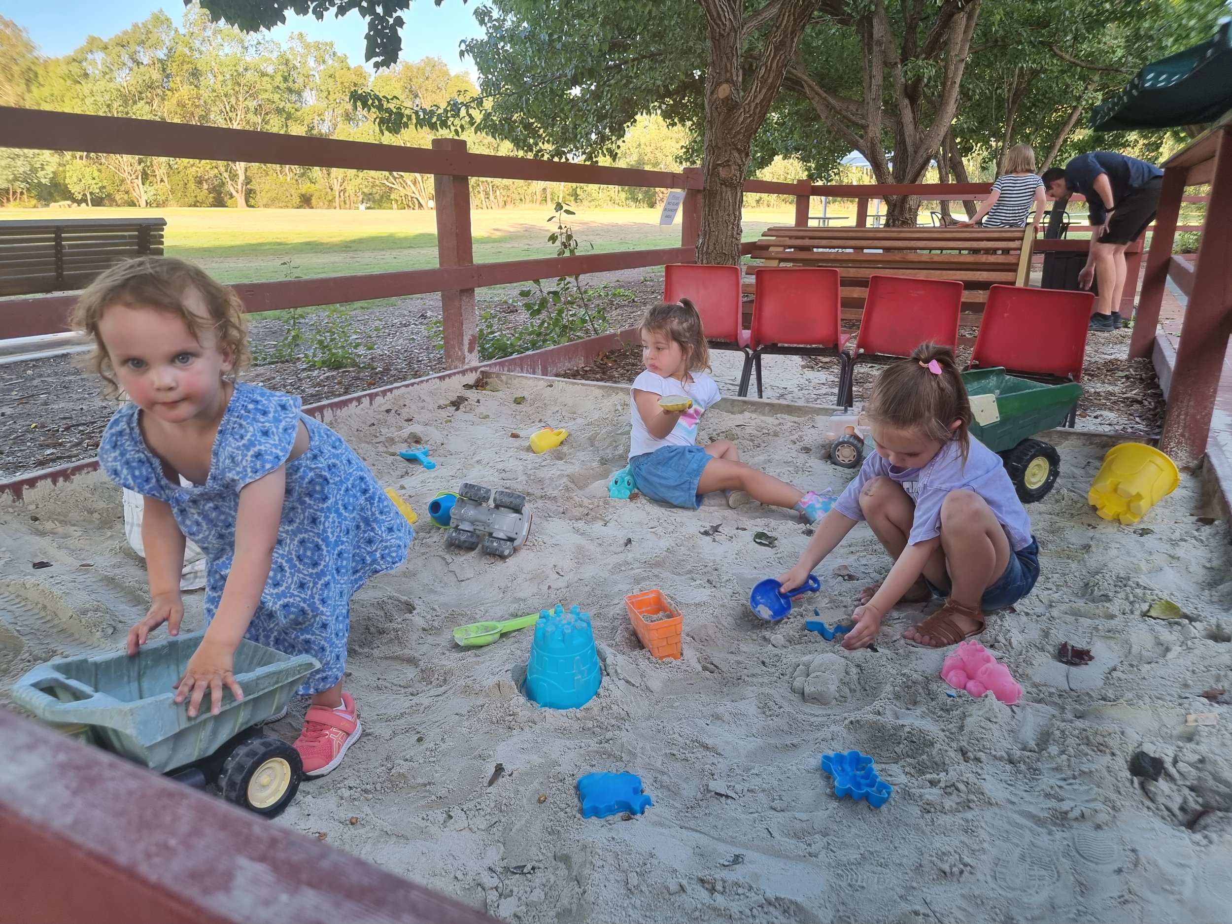 Children playing and digging in a sandbox at a park with trees and benches in the background.