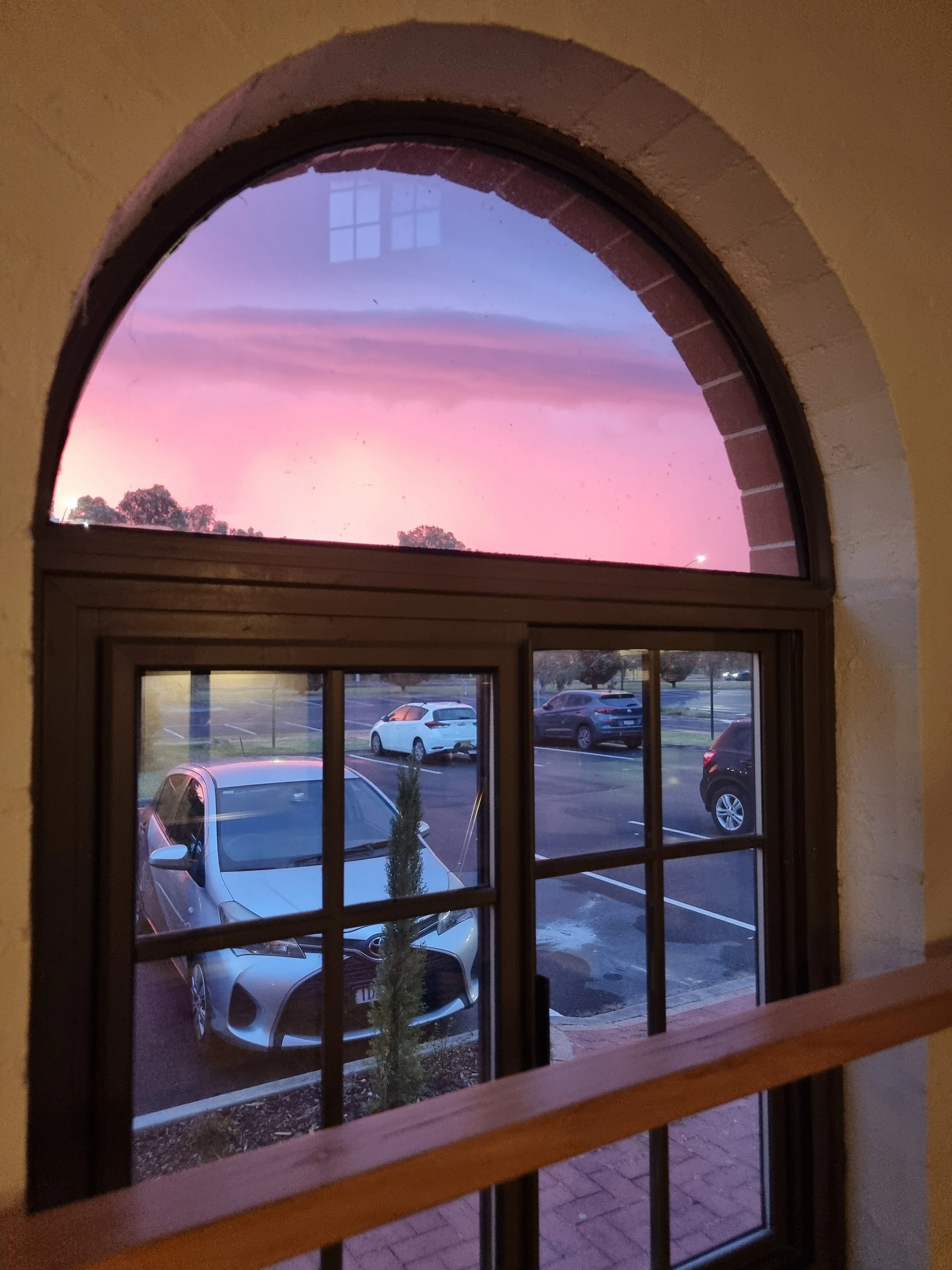 View through a window with an arched top and grid panes showing parked cars and a sunset sky with pink and purple hues.
