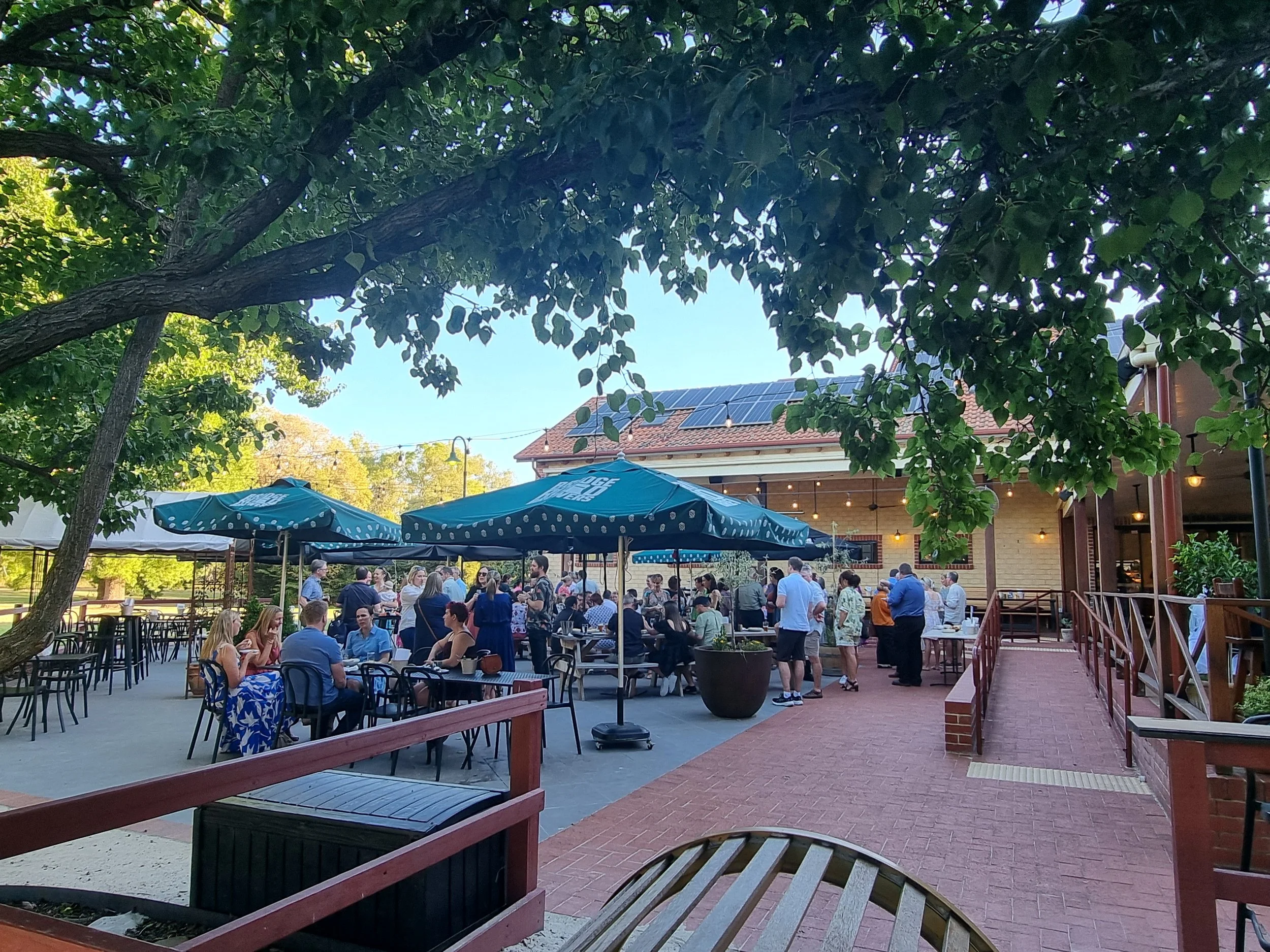 People dining outdoors at a restaurant with large blue umbrellas, under a leafy tree, on a brick and concrete patio with a building in the background.