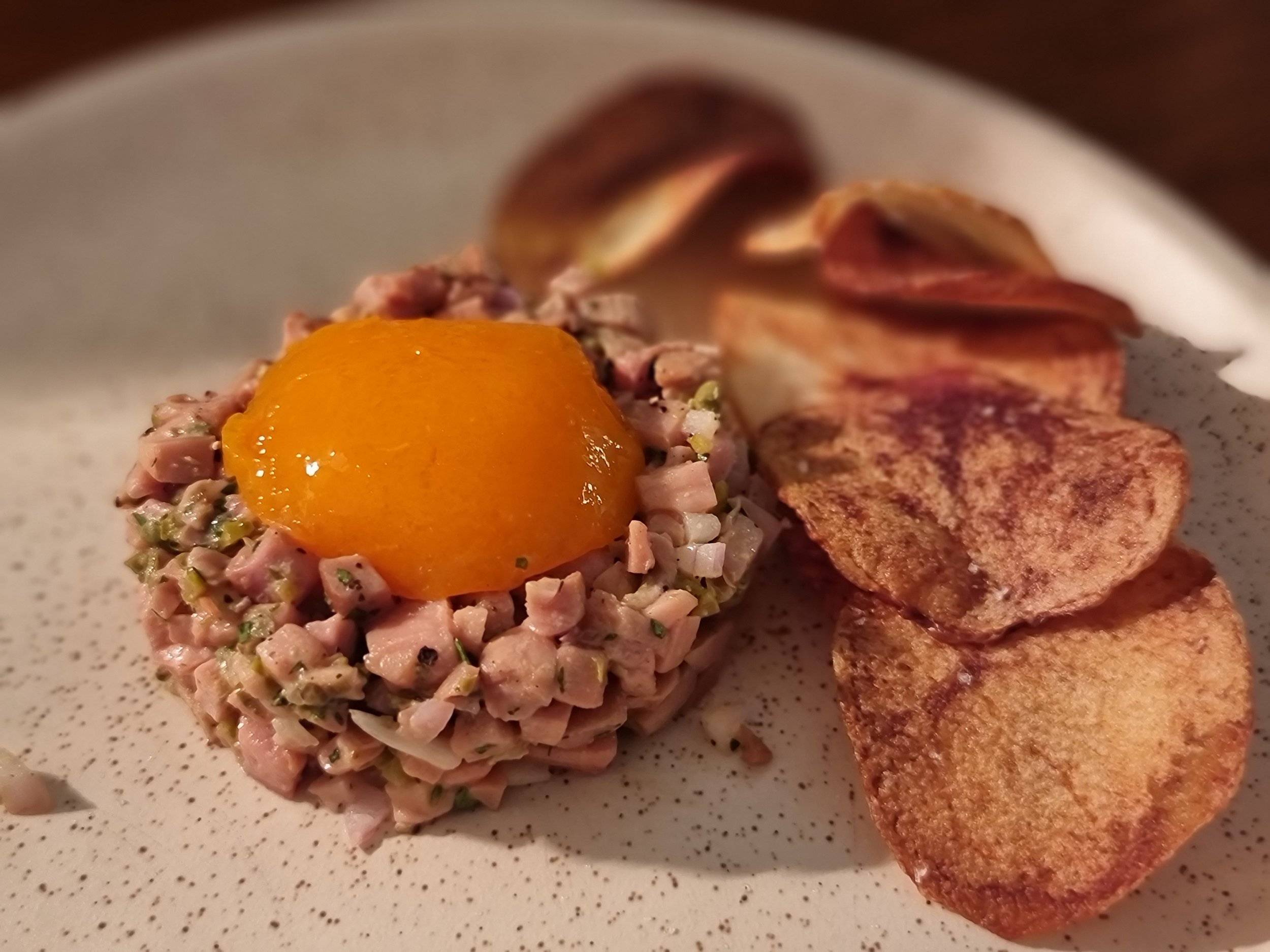 Beef tartare topped with a raw egg yolk, served with potato chips on a speckled beige plate.