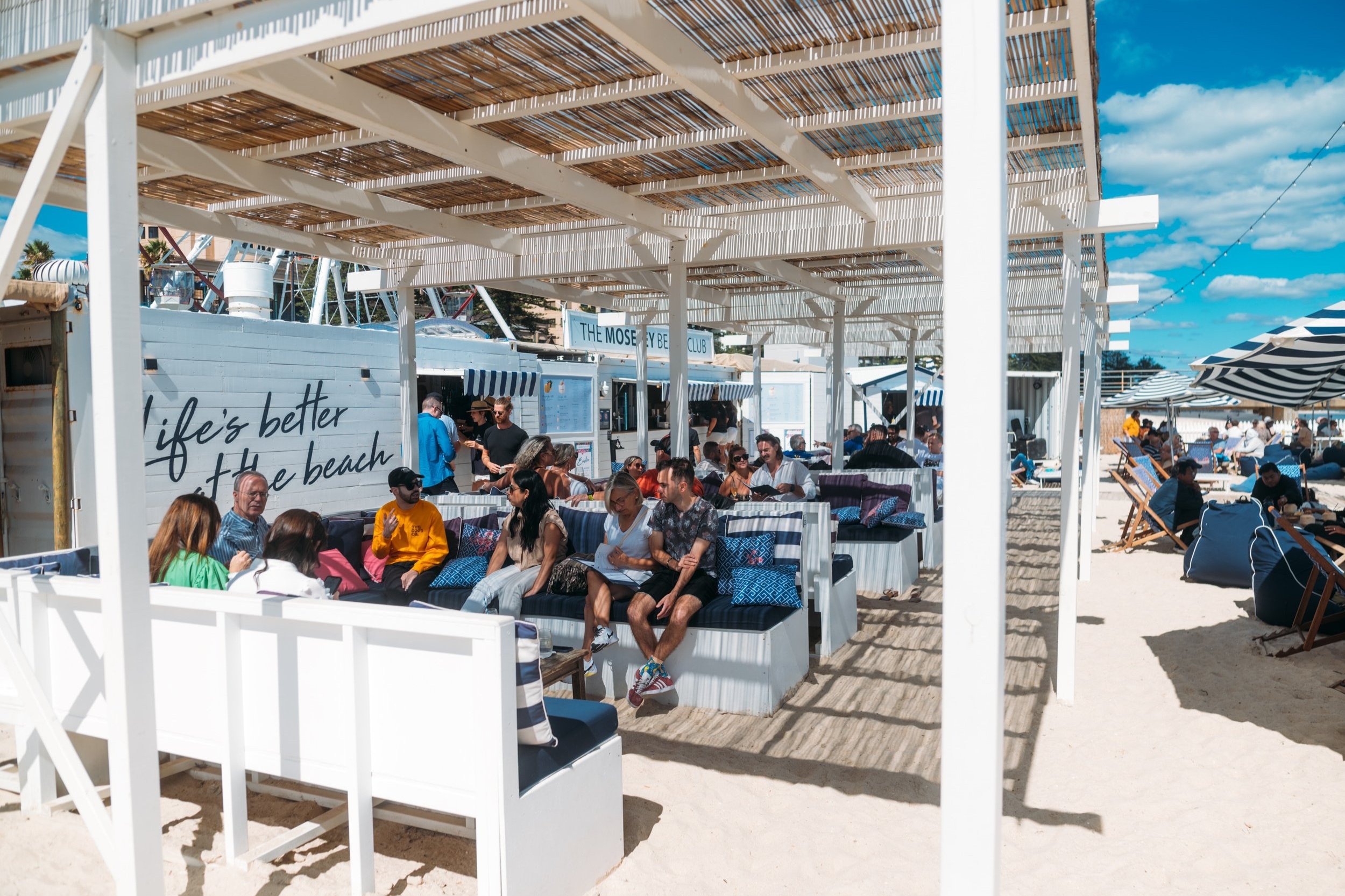People sitting and chatting at a beachside restaurant with white and blue decor, under a wooden pergola, on a sunny day with blue skies and scattered clouds.
