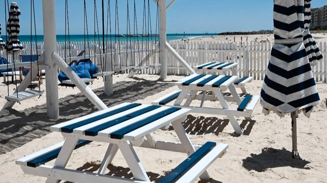 Beach chairs and umbrellas on sandy beach near white picket fence, with ocean and buildings in the background.