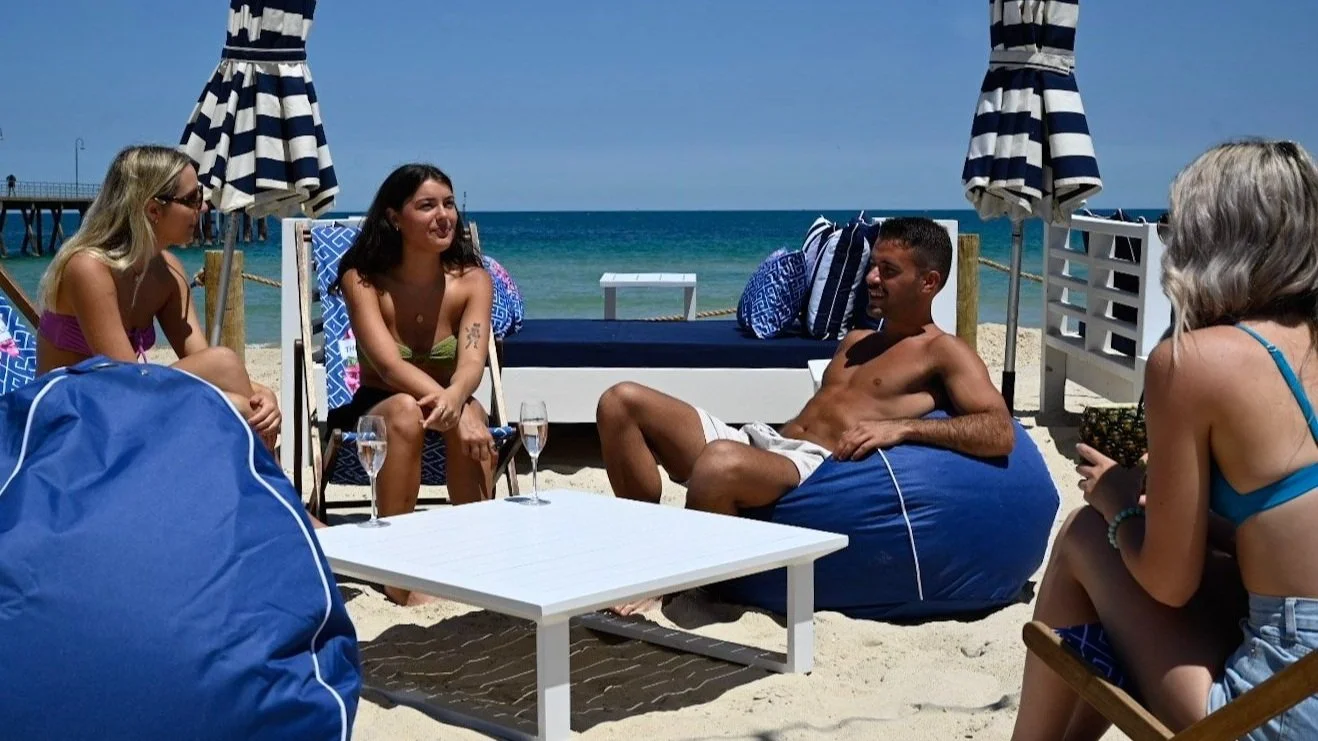 Group of people relaxing and chatting on a beach, sitting on bean bags and chairs under large striped umbrellas with the ocean in the background.