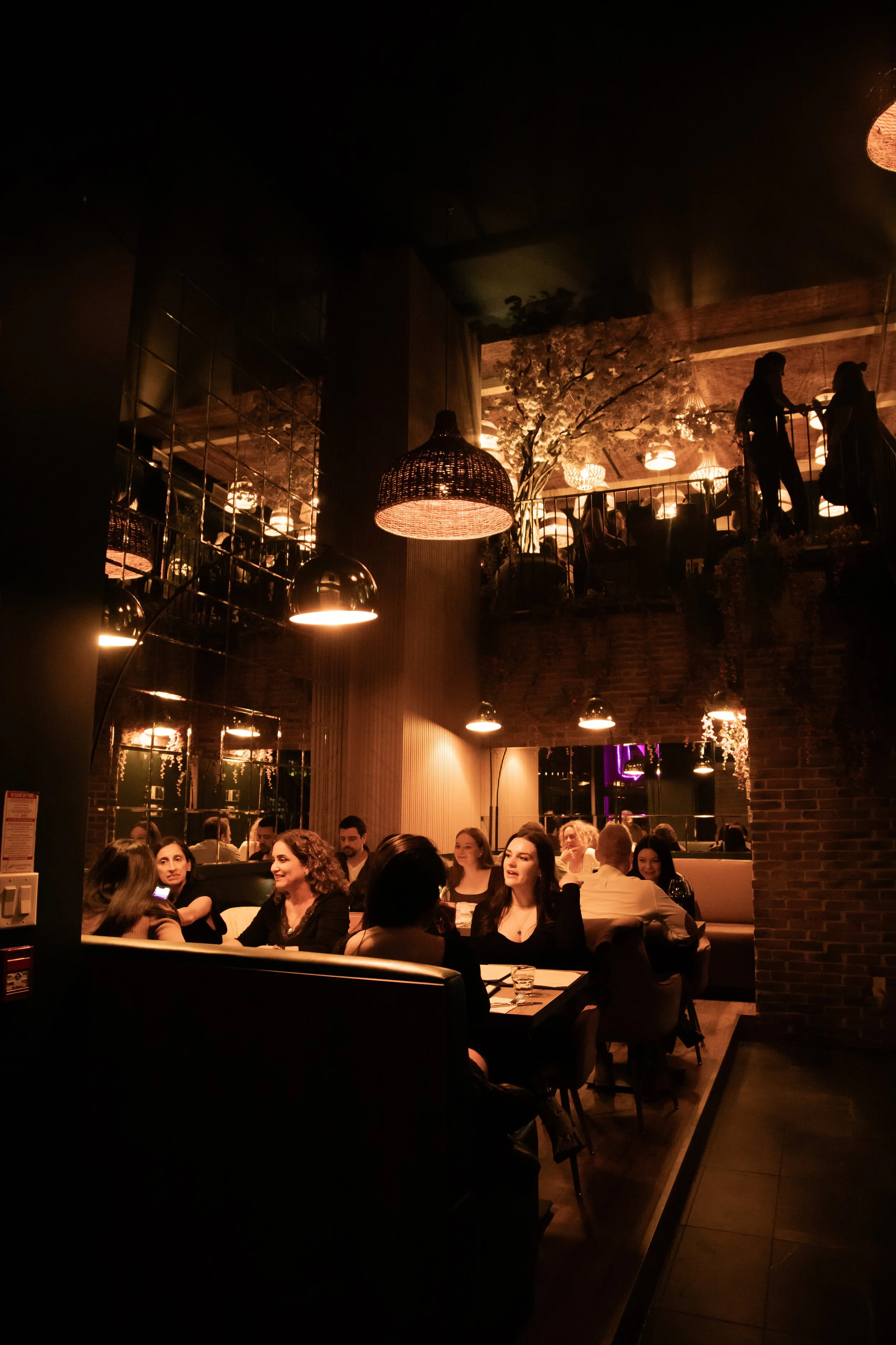People dining in a cozy restaurant with warm lighting, dark walls, and decorative elements like hanging lamps and plants, with a second-floor balcony visible.