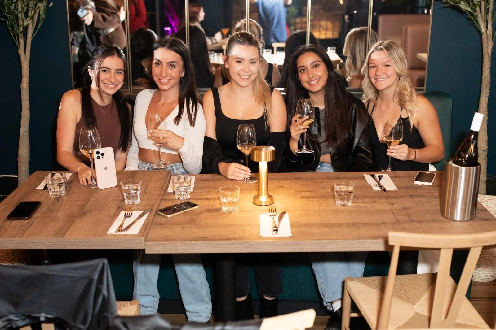 Five women sitting at a restaurant table, smiling and holding wine glasses, with a mirror and other patrons in the background.