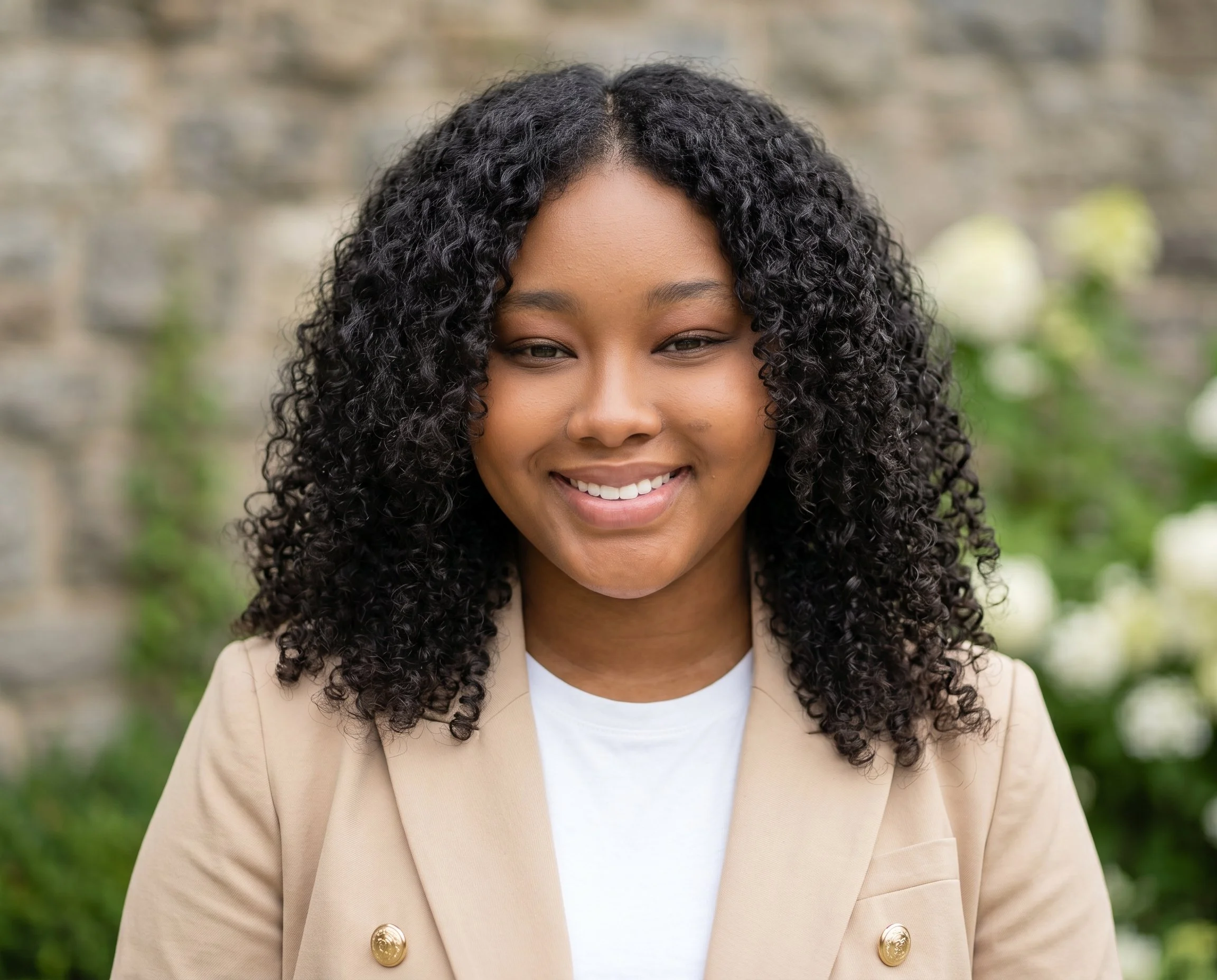 A woman with curly black hair, smiling, wearing a beige blazer with gold buttons and a white top, outdoors with blurred greenery and stone wall background.