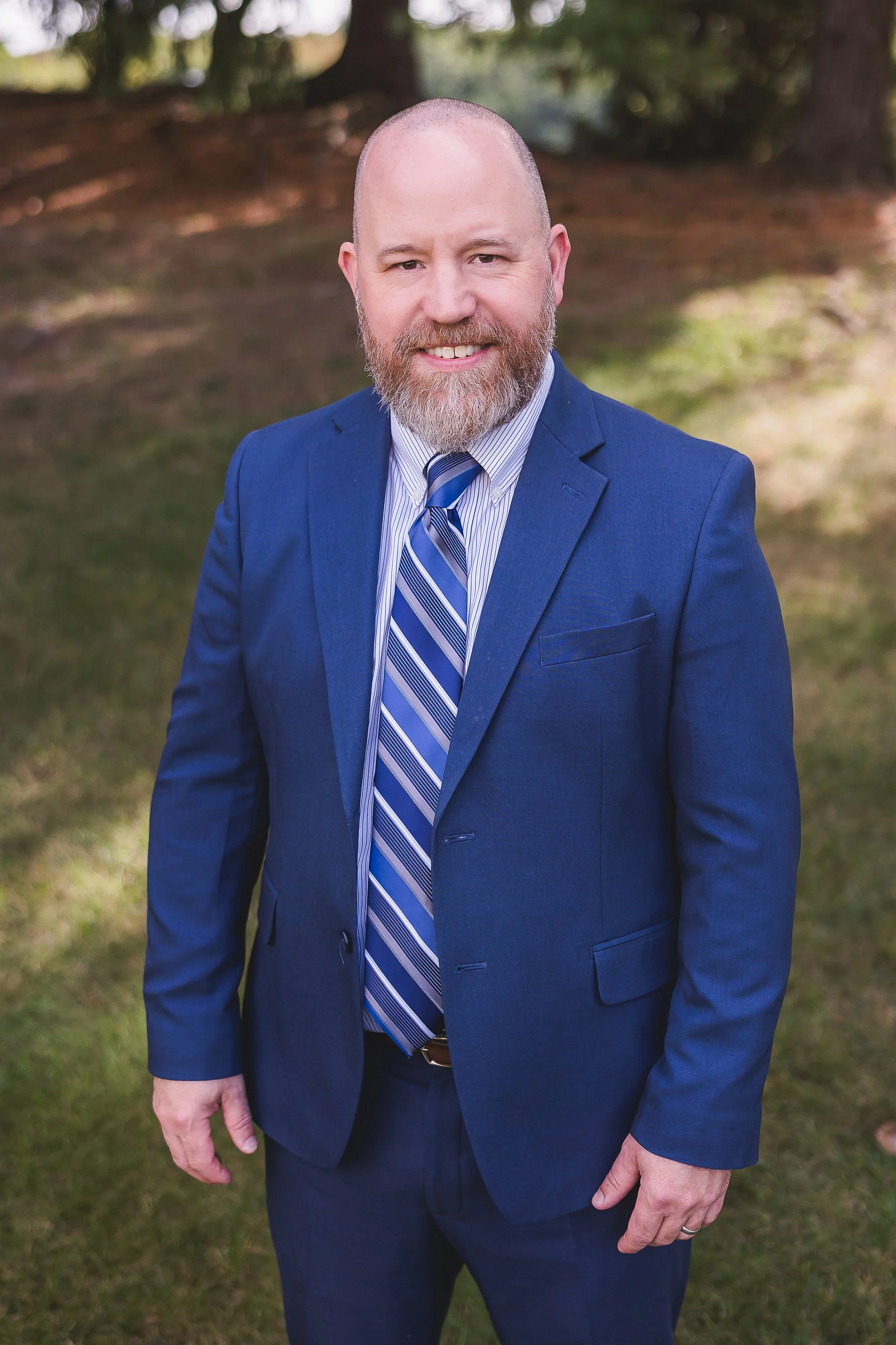Bald man with a beard in a blue suit and plaid tie standing indoors next to a plant