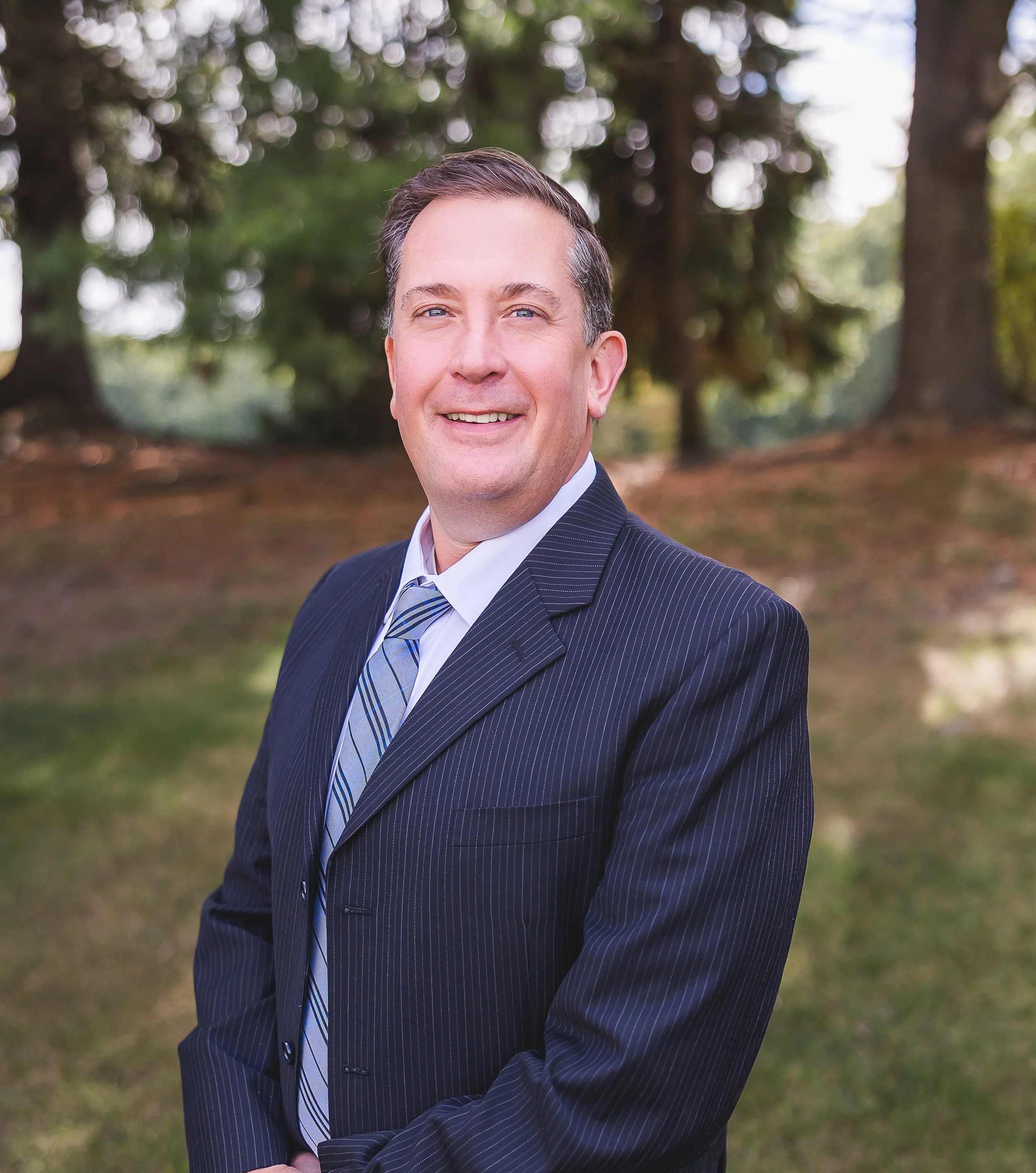 A man in a dark suit with a blue-striped tie, smiling outdoors with a blurred background of trees and greenery.