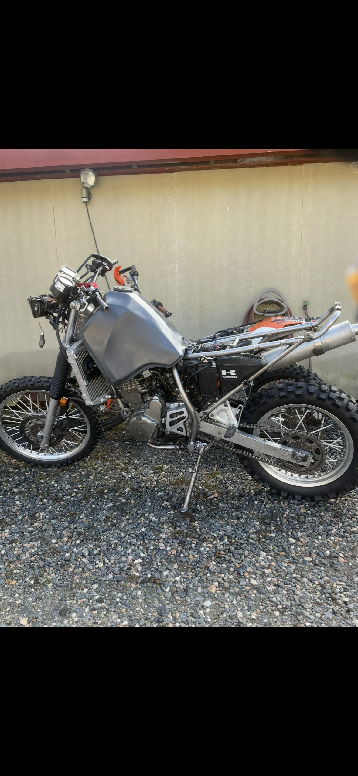A partially assembled off-road motorcycle with knobby tires, a black Kawasaki logo on the side, and an exposed frame and engine, parked on a gravel surface next to a yellow wall with a garden hose.