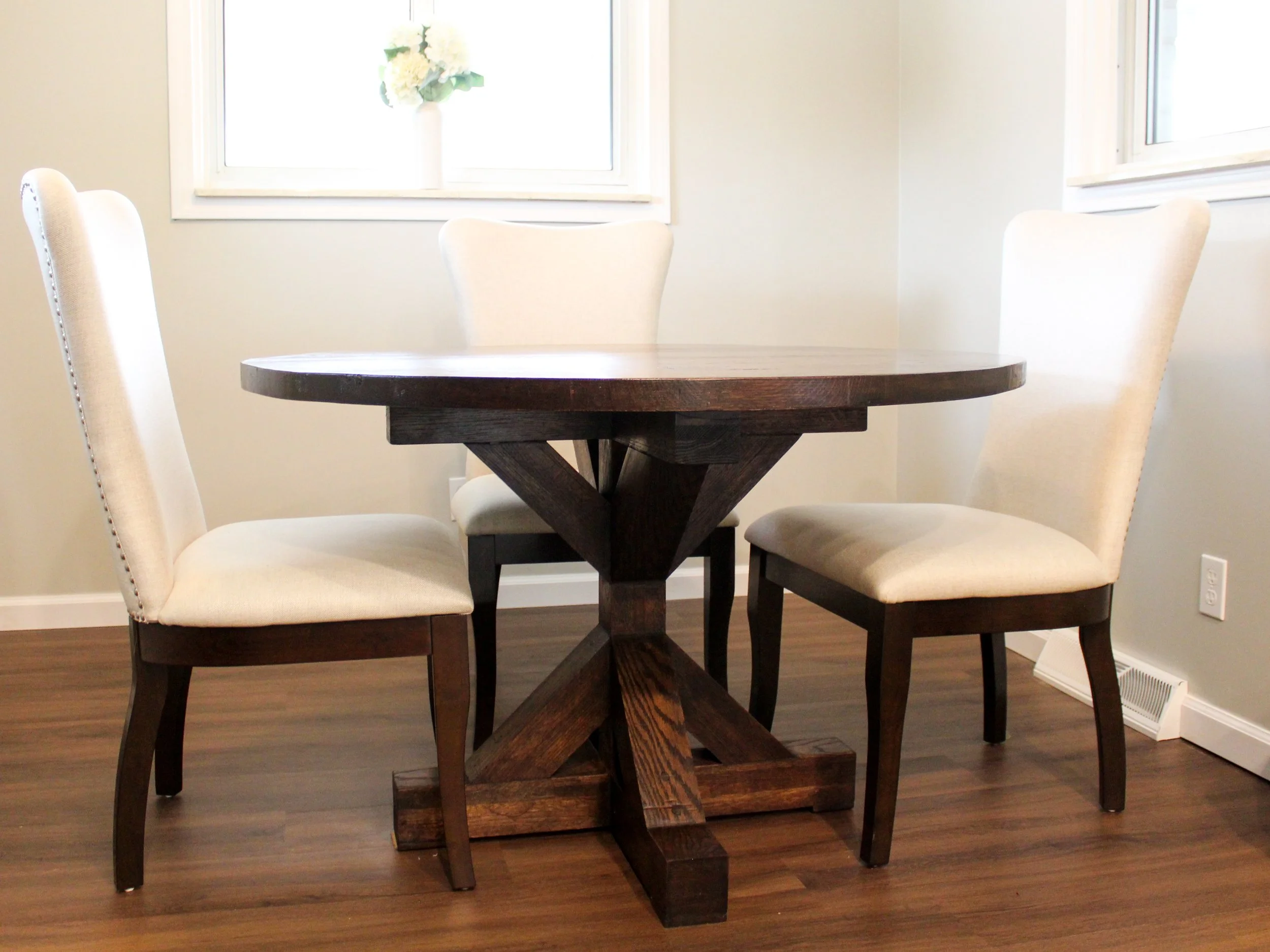 A round wooden dining table with a white vase holding white flowers, surrounded by four beige upholstered chairs with nailhead trim, in a room with beige walls, wood flooring, and two windows.