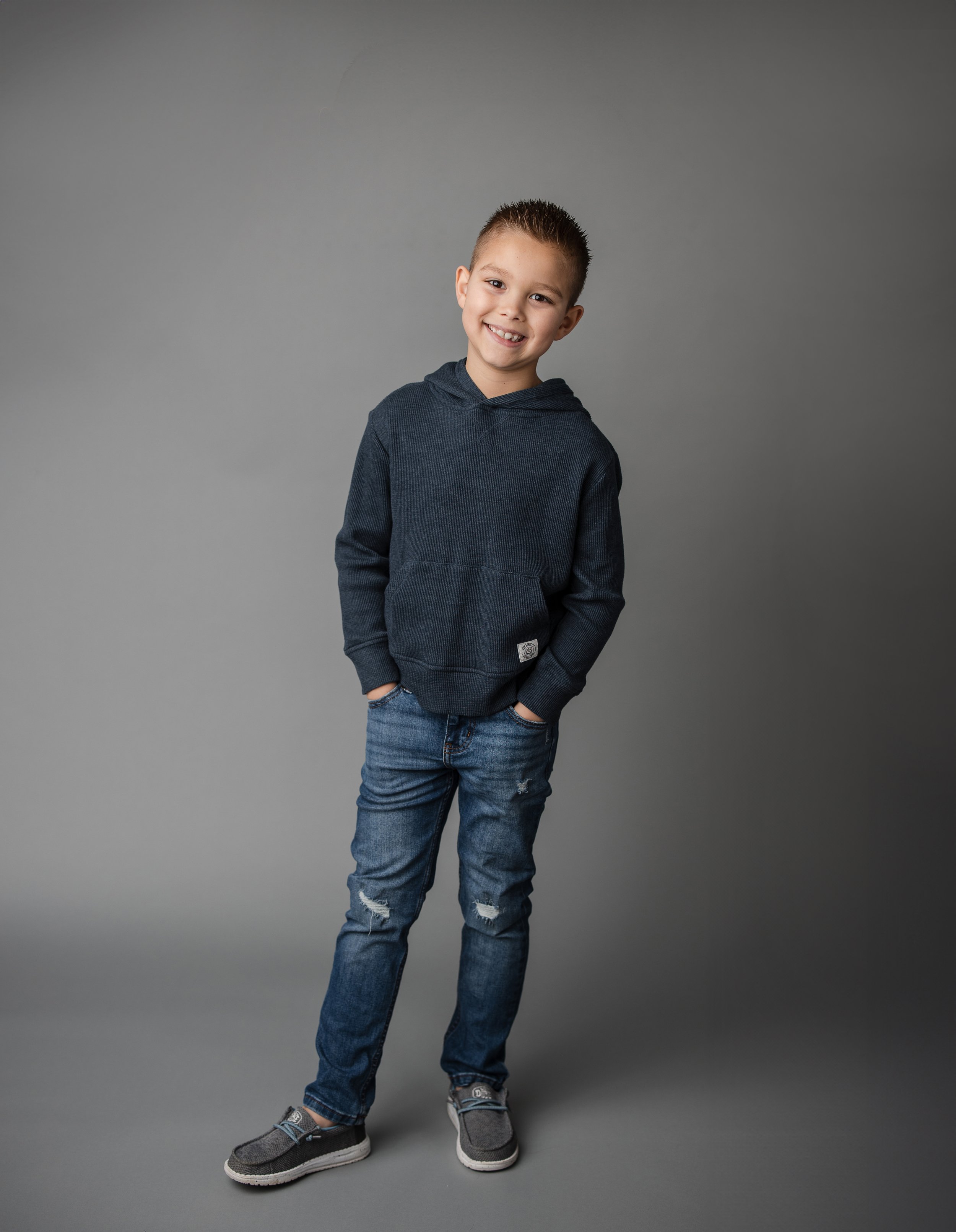 Smiling boy in dark hoodie and jeans against gray backdrop