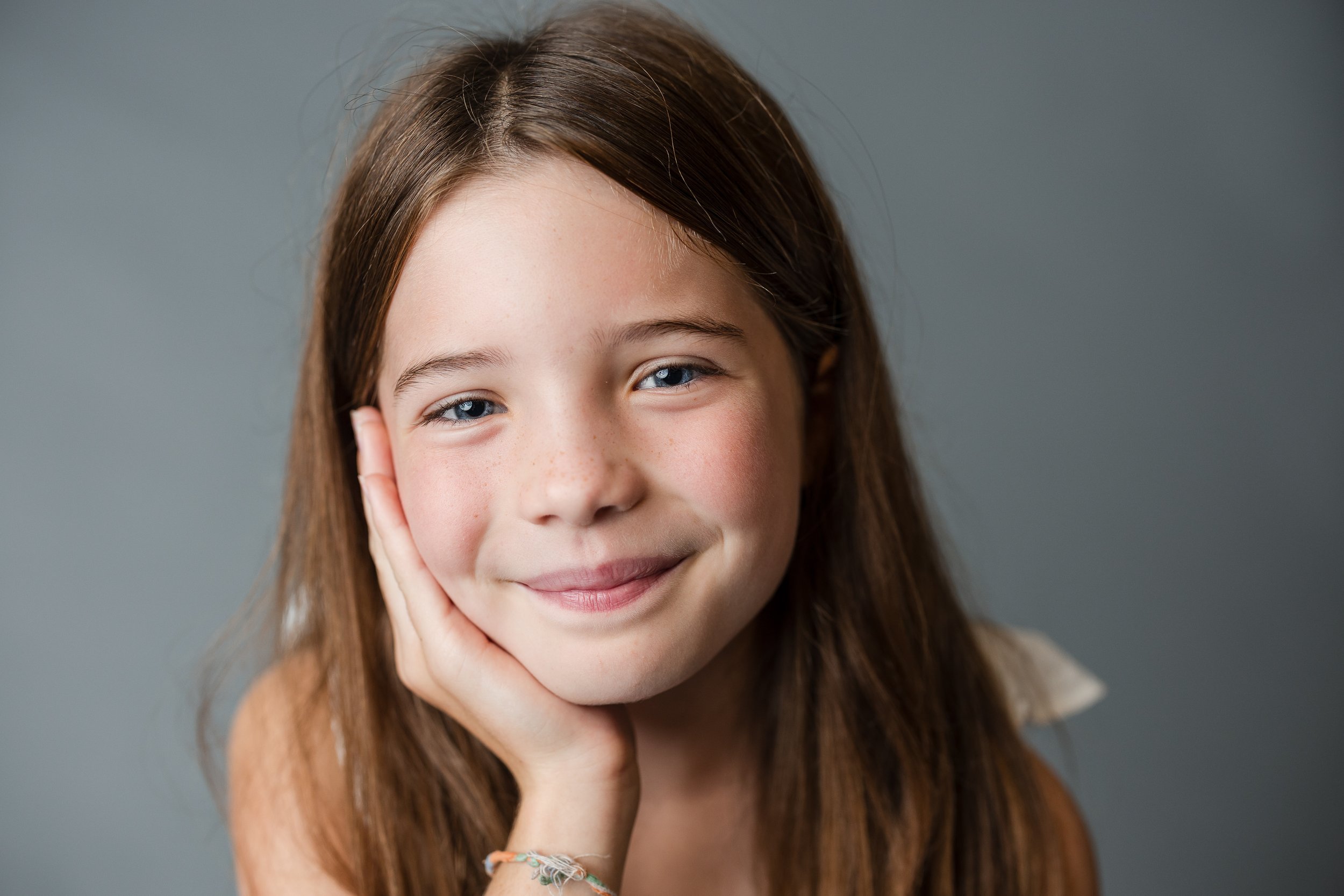 Young girl smiling with hand on her face, long brown hair, gray background.