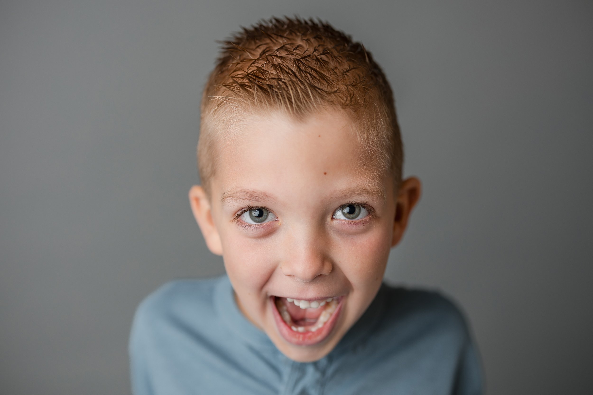 Close-up of a young child with short hair smiling excitedly, wearing a light blue shirt against a gray background.