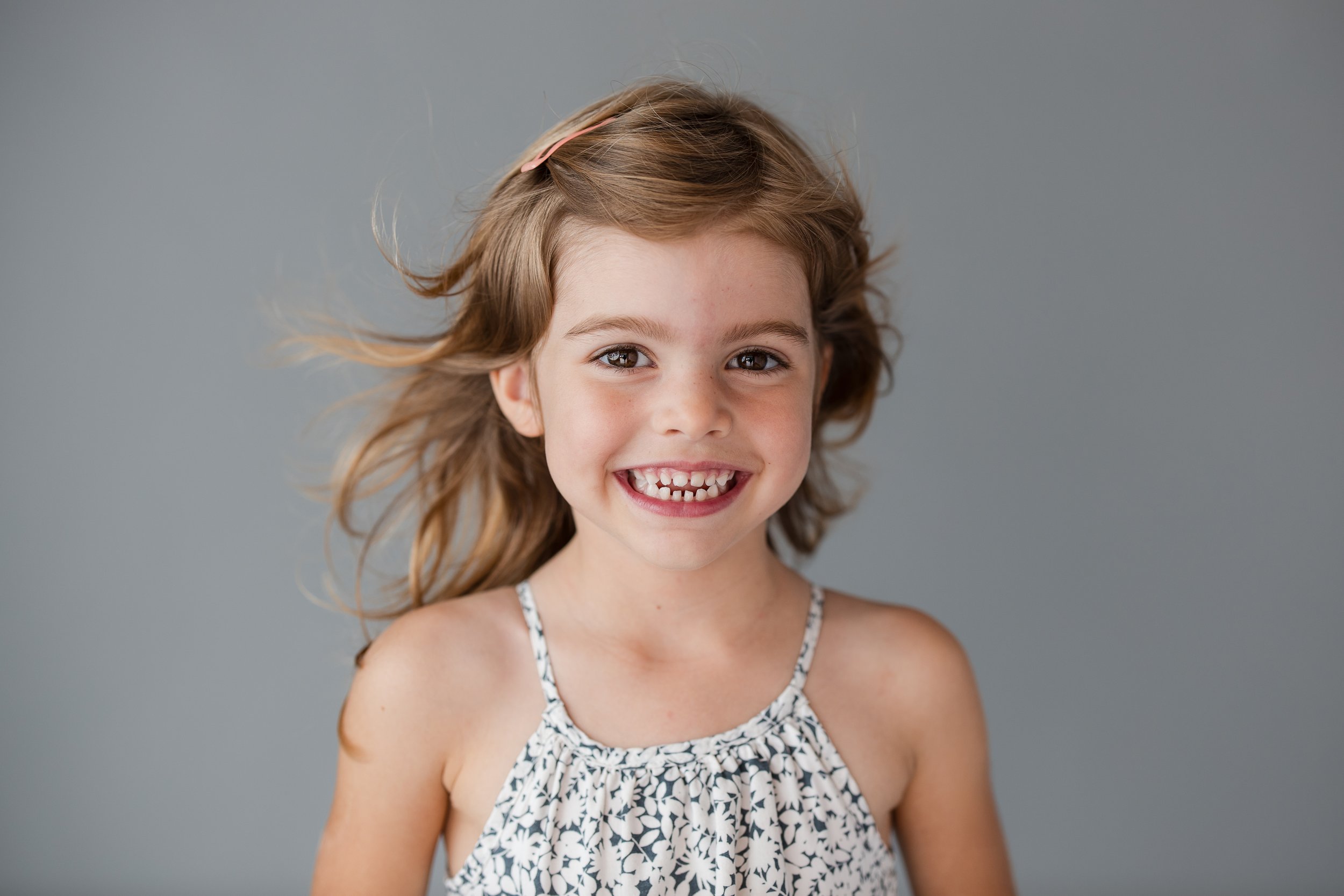 Smiling young girl with long hair wearing a patterned dress against a plain background.