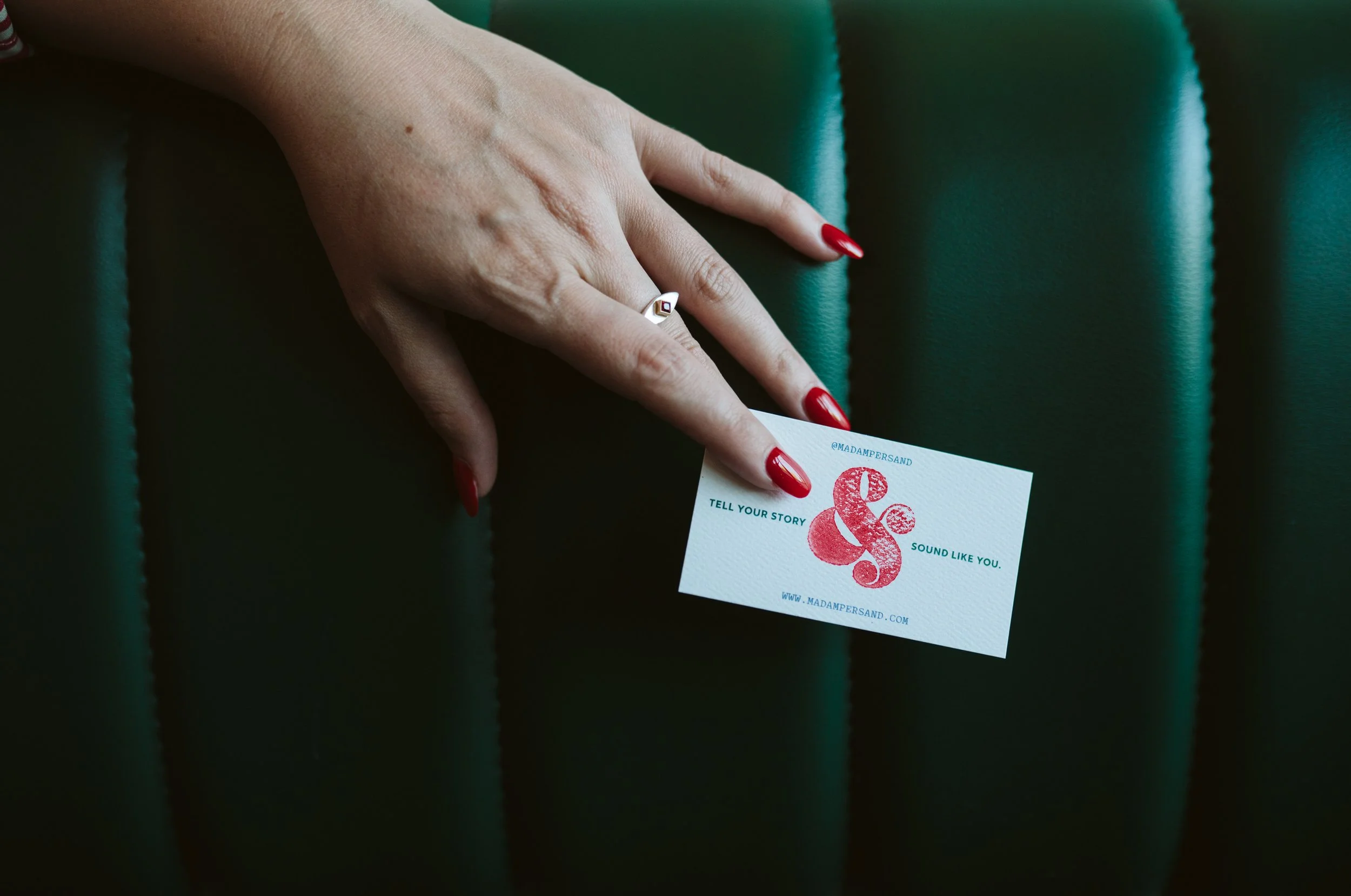 Meg's business card, held lightly between manicured nails against a green leather backdrop.