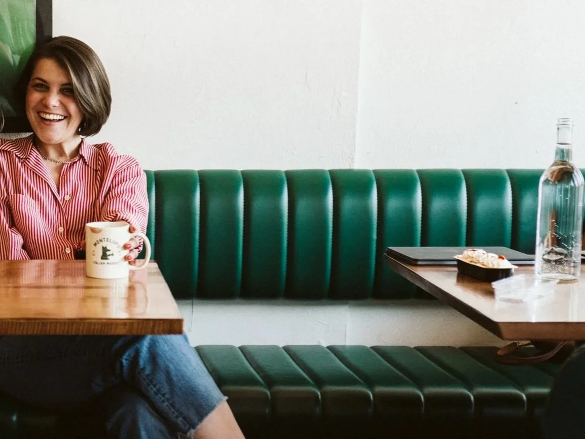 Meg laughs, seated in a green leather booth with a coffee in hand.