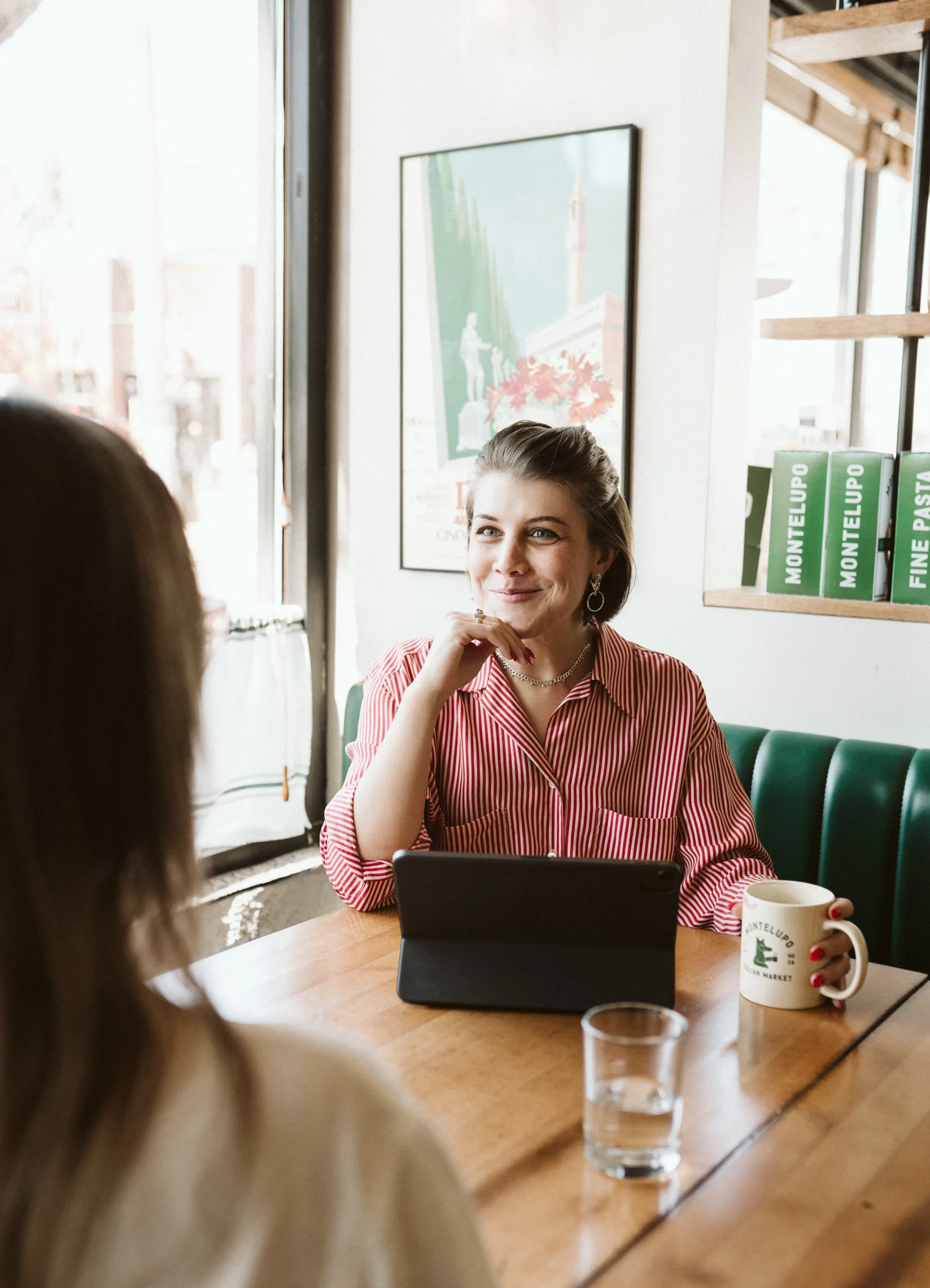 A woman sitting at a wooden table in a cafe, smiling and looking at someone across from her. She has short dark hair, wears earrings, a red and white striped shirt, and has a mug and a tablet in front of her. There are shelves with books and a poster on the wall behind her.