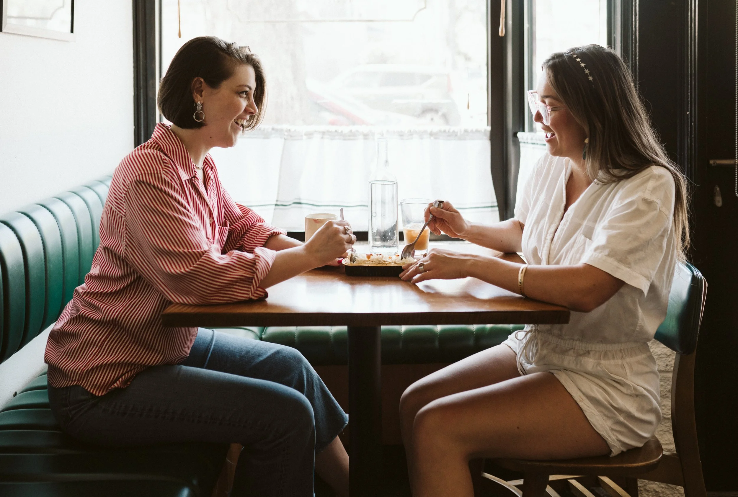 Two women sit across from each other in a cafe, smiling and engaged in conversation. One woman has short brown hair, and the other has long dark hair with a star headband, wearing glasses. They are sharing a meal on a wooden table near a window.