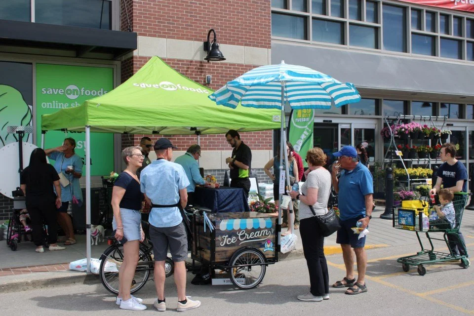 People gather outside Save-On-Foods around an ice cream cart with a striped umbrella, enjoying treats on a sunny day.