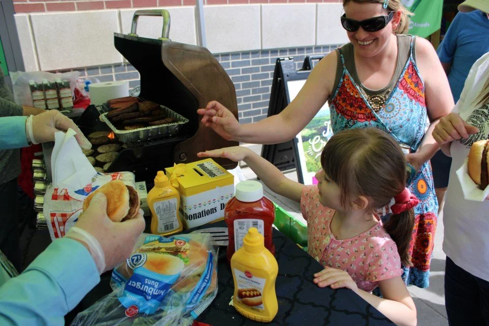 A young girl reaches for condiments at a barbecue table while a woman beside her smiles; a person wearing gloves prepares burgers near a grill in the background.