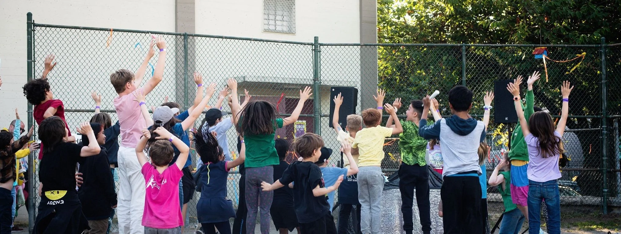 Children outdoors near a chain-link fence, raising their arms and cheering together during an activity.s standing near a chain-link fence, raising their hands in the air.