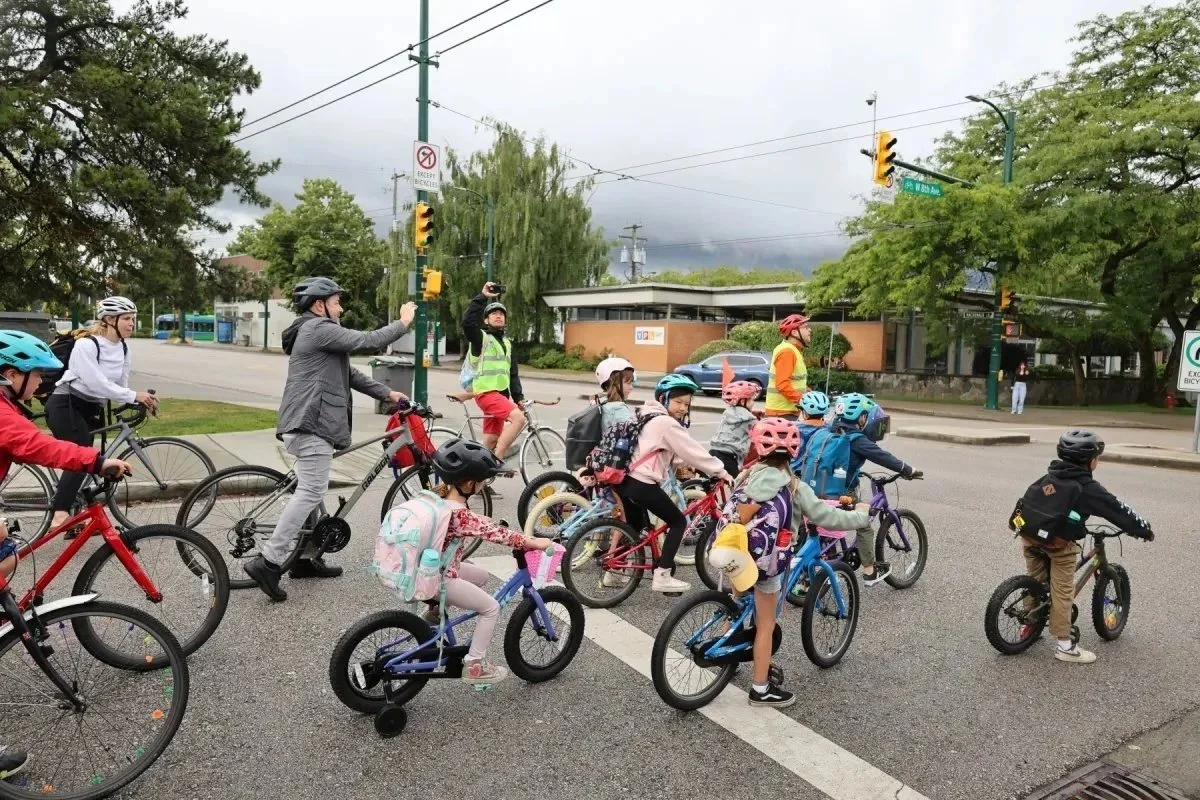 A group of elementary school children and adults ride bicycles together through a city intersection during a bike-to-school event. The children wear colorful helmets and backpacks while crossing the street under cloudy skies.