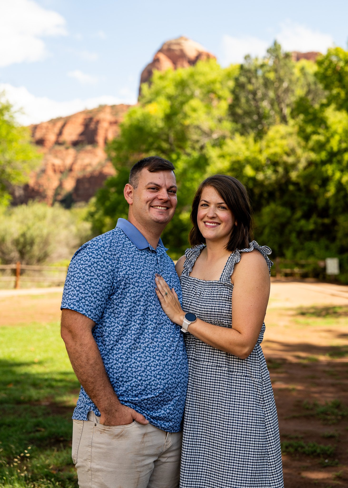 A smiling couple standing outdoors in front of trees and red rock formations, with the woman placing her hand on the man's chest, both looking at the camera.
