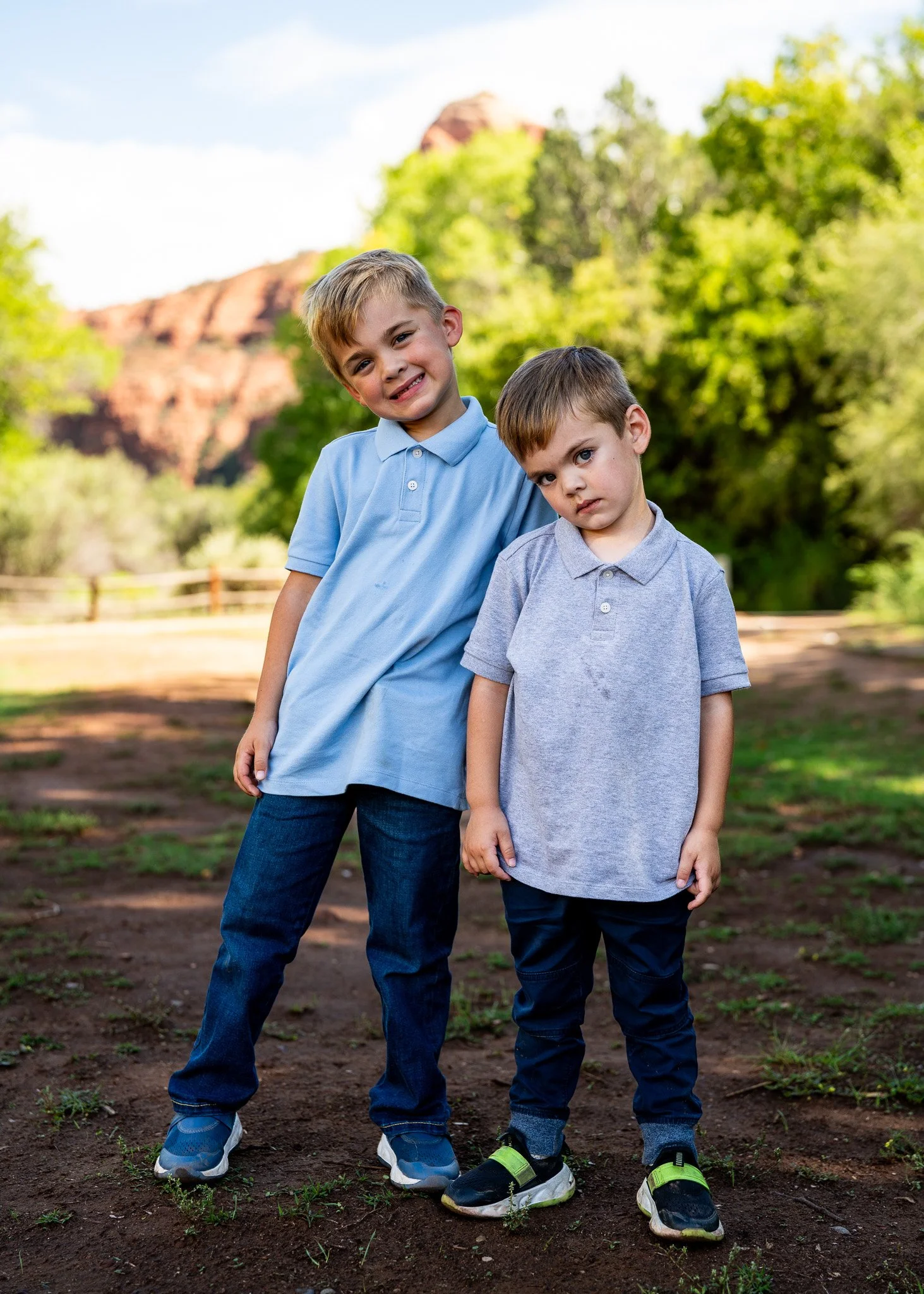 Two young boys standing outdoors on a dirt patch with green trees and rocky hills in the background. One boy is leaning his head on the other's shoulder, both wearing casual polo shirts and jeans, with the older boy smiling and the younger boy lookin