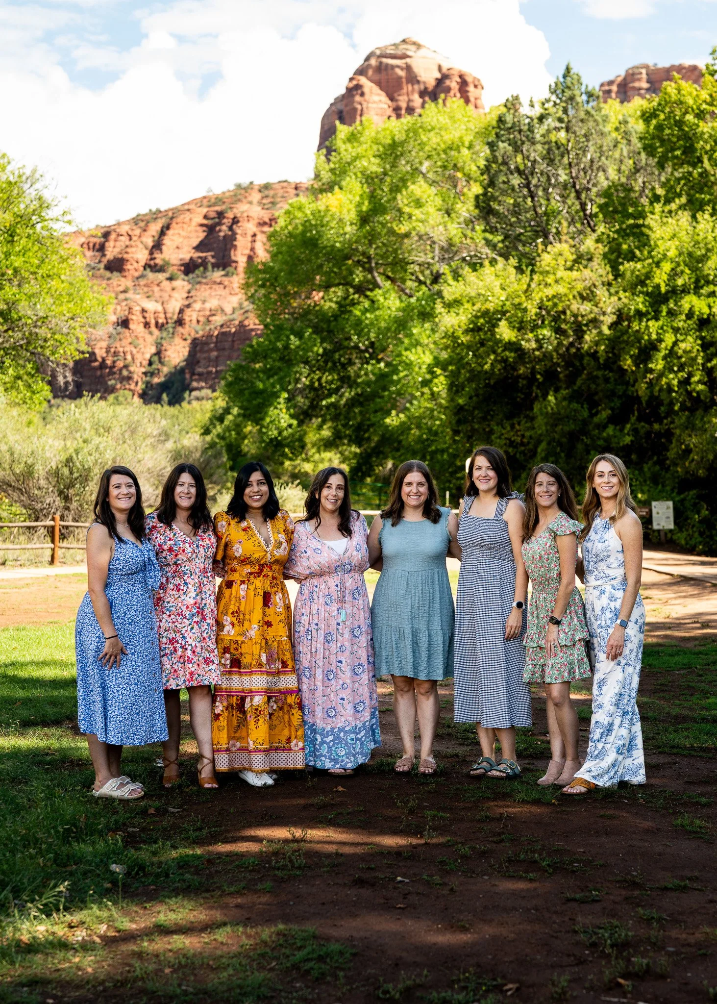 Group of nine women standing outdoors in front of trees and red rock formations, smiling and posing for the photo.