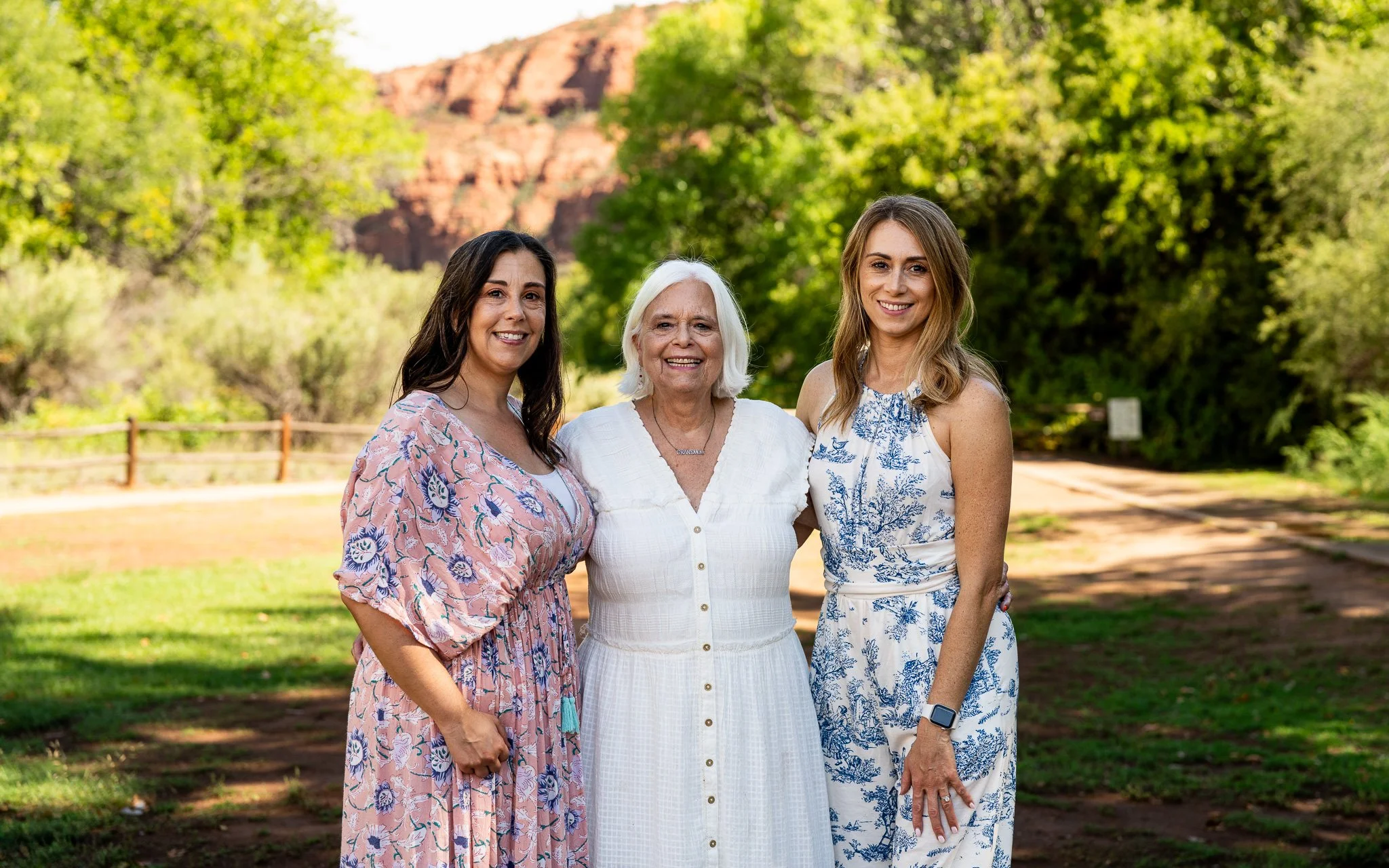 Three women standing outdoors smiling with trees and a dirt path behind them.