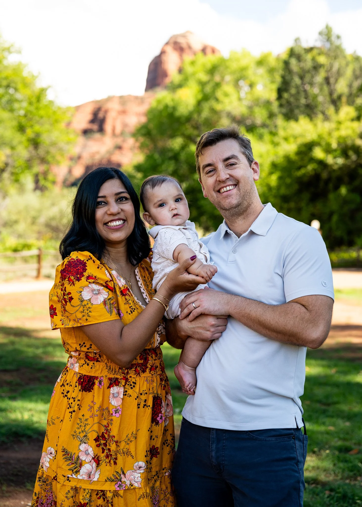 A family of three smiling outdoors in front of a scenic background with green trees and a large rock formation. The woman is wearing a yellow floral dress, the man a white polo shirt, and they are holding a baby.