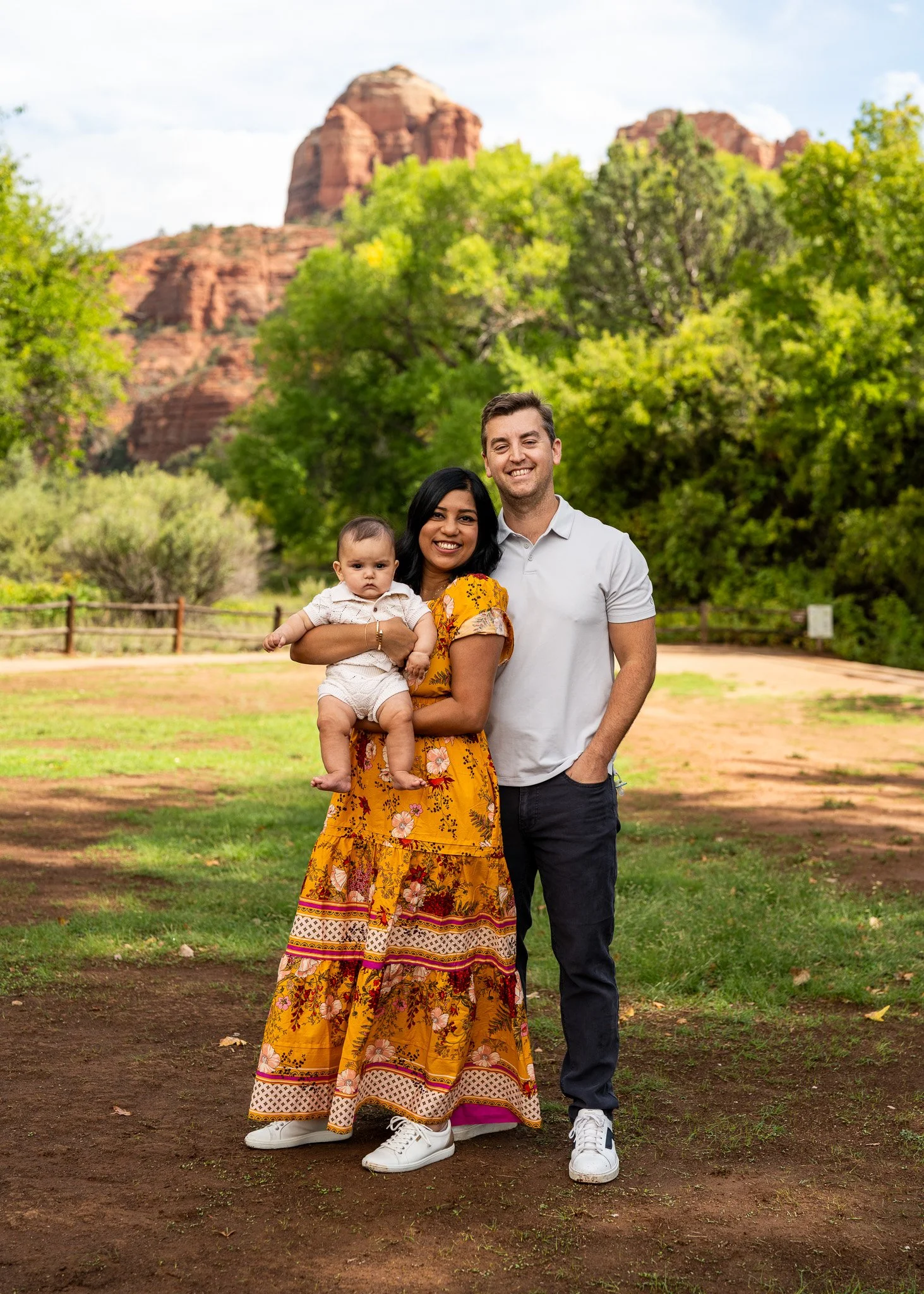 A happy family of three standing outdoors in a park with green trees and red rock formations in the background. The woman is holding a baby girl, and both are smiling at the camera.