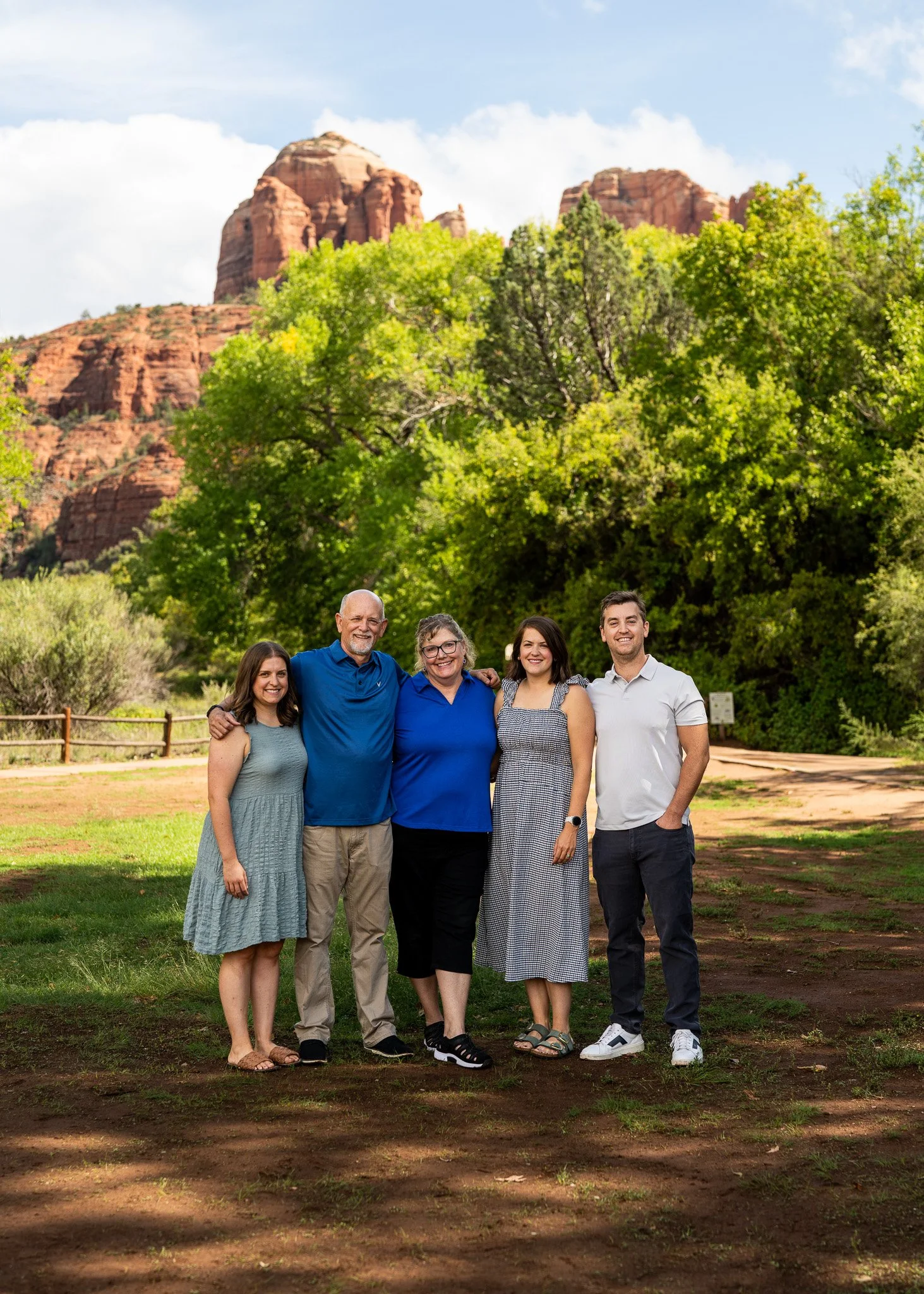 A family portrait outdoors with red rock cliffs and green trees in the background, including three women and two men standing close together and smiling.