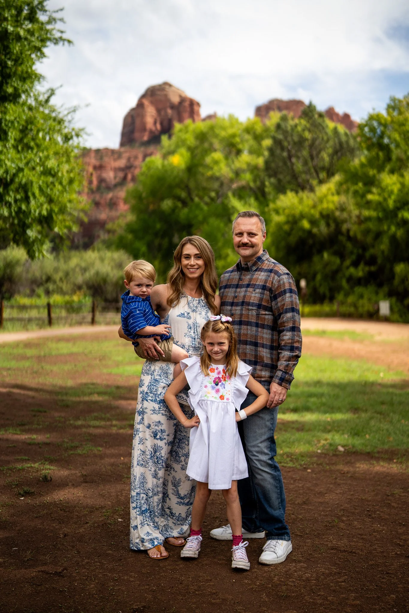 A family of four posing outdoors in a park with trees and red rock formations in the background, under a partly cloudy sky.