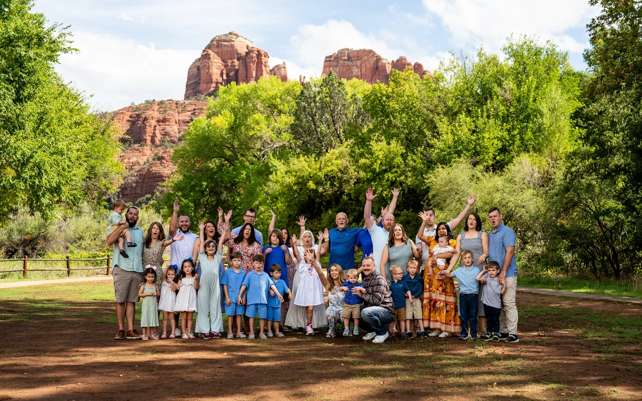 Large group of people, including adults and children, gathered outdoors in a park with green trees and red rock formations in the background, posing for a group photo with some people raising their hands and smiling.