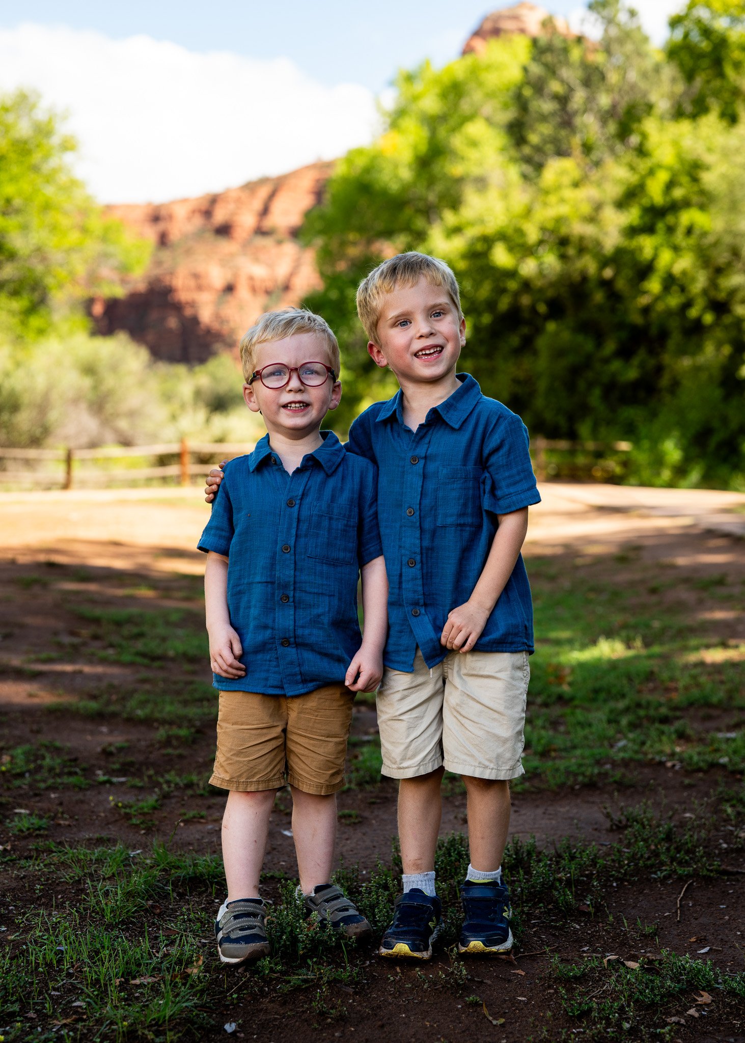 Two young boys standing outdoors in a park with trees and red rock formations in the background. They are hugging each other and smiling, dressed in matching blue shirts and shorts.