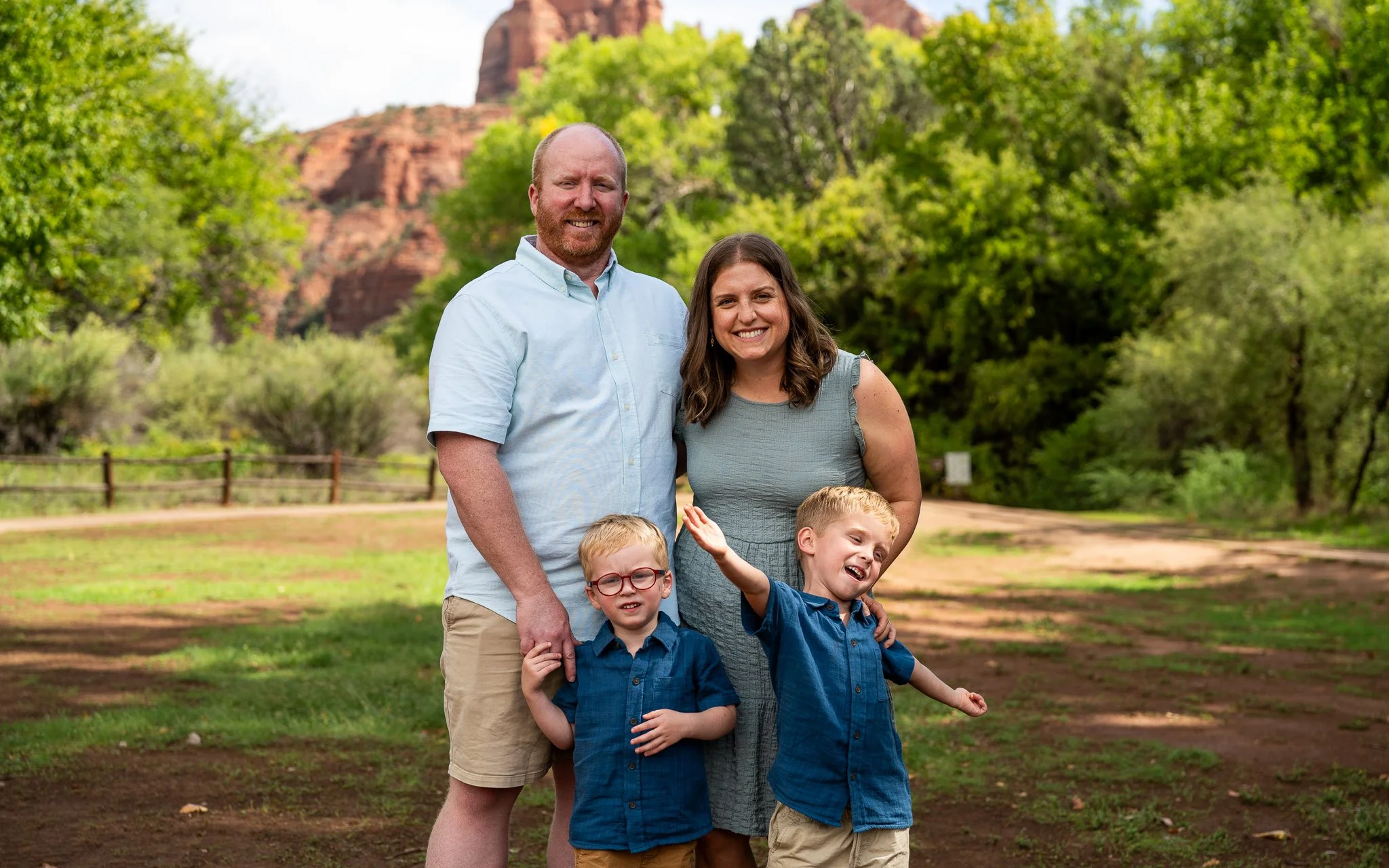A family of four standing outdoors in a park with trees and a rocky hill in the background. The father has a beard and is wearing a light blue shirt and khaki shorts. The mother has shoulder-length brown hair and is wearing a gray dress. Their two yo