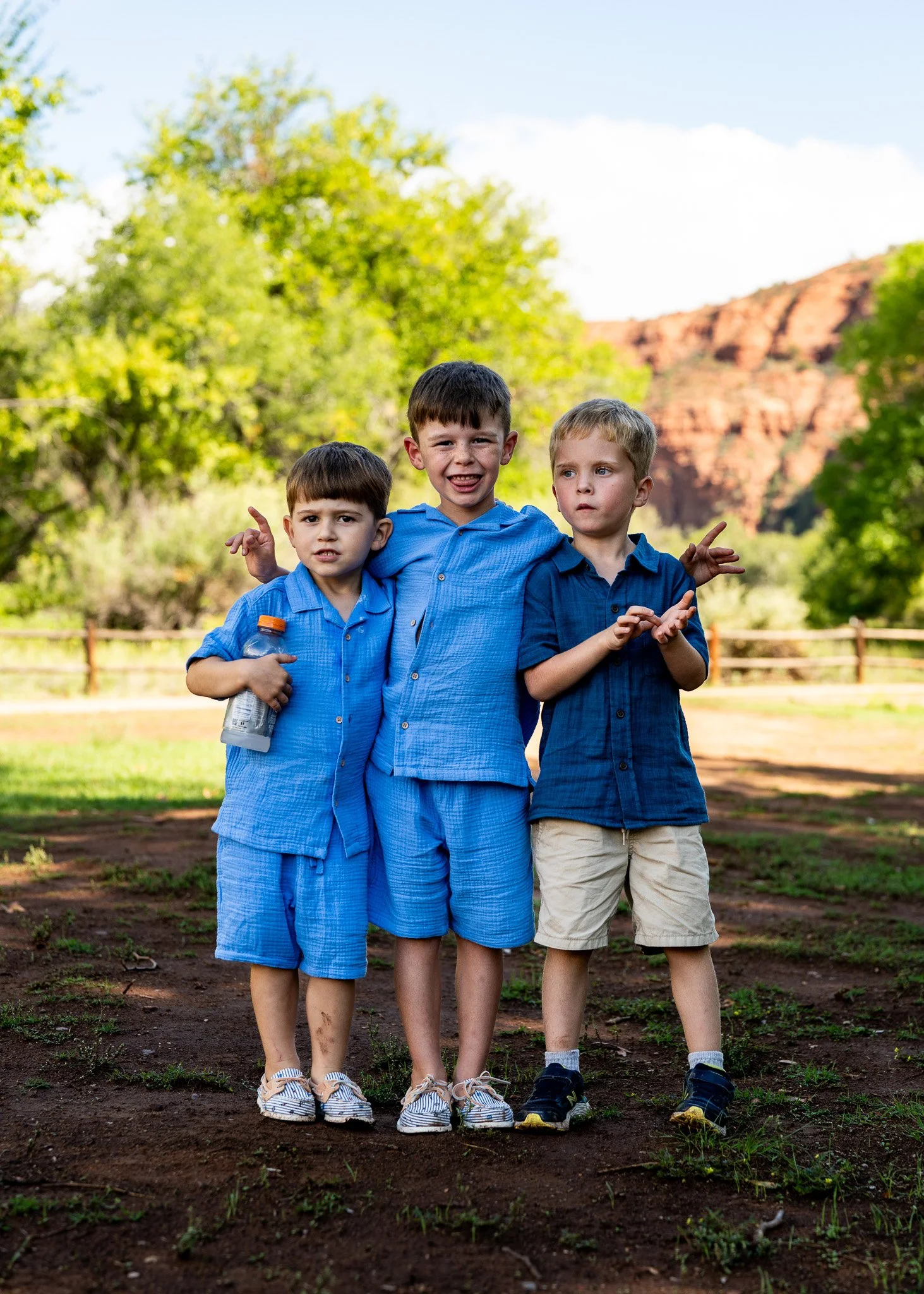 Three young boys standing outdoors on dirt ground with trees and a rocky hill in the background, wearing casual blue shirts and shorts.