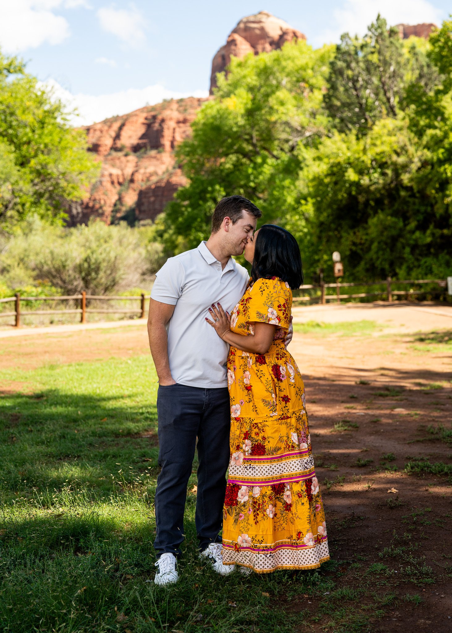 A couple kissing outdoors in a park with green trees and red rock formations in the background, during a sunny day.