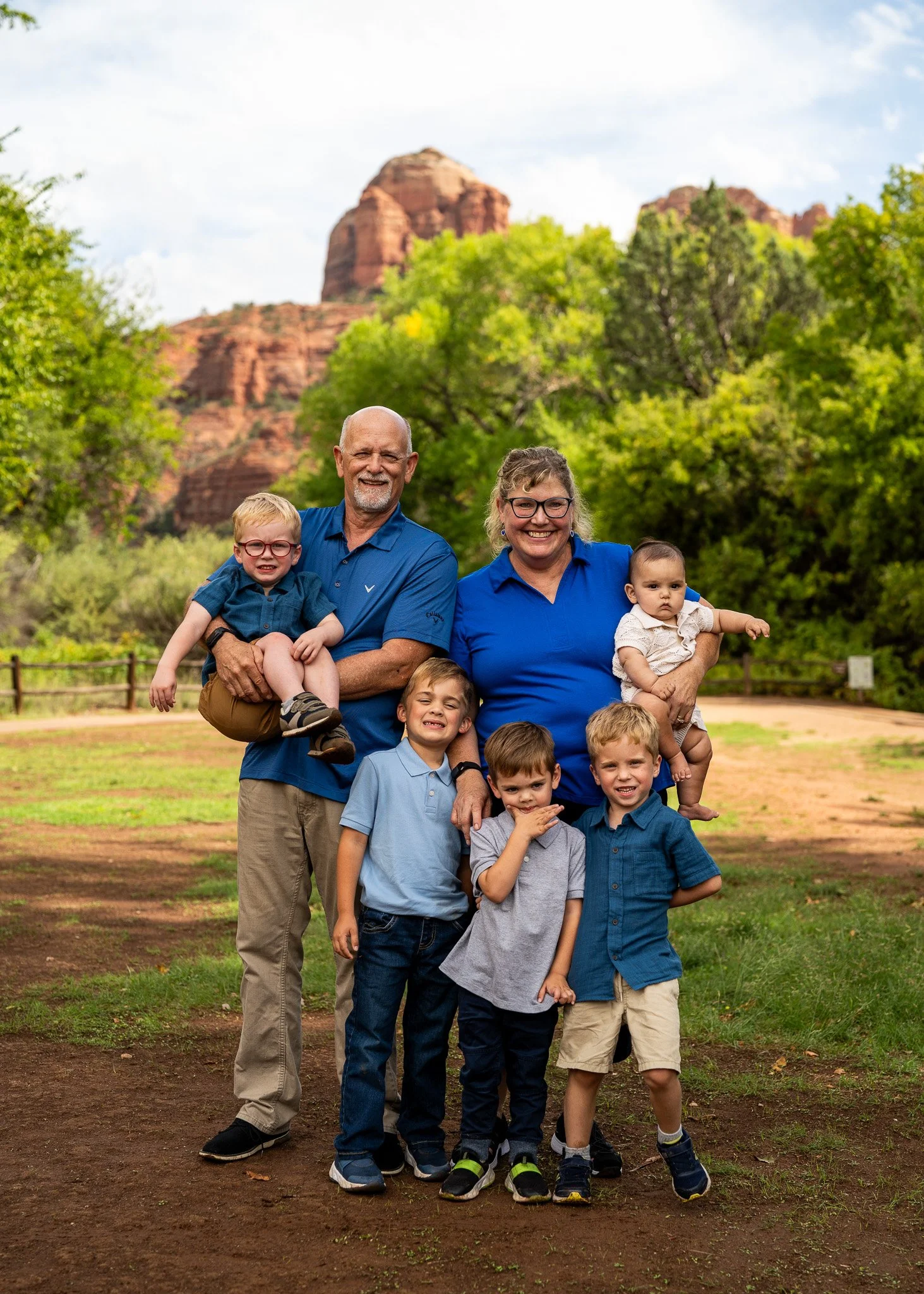 A family of eight posing outdoors in front of red rock formations and green trees, with a partly cloudy sky.