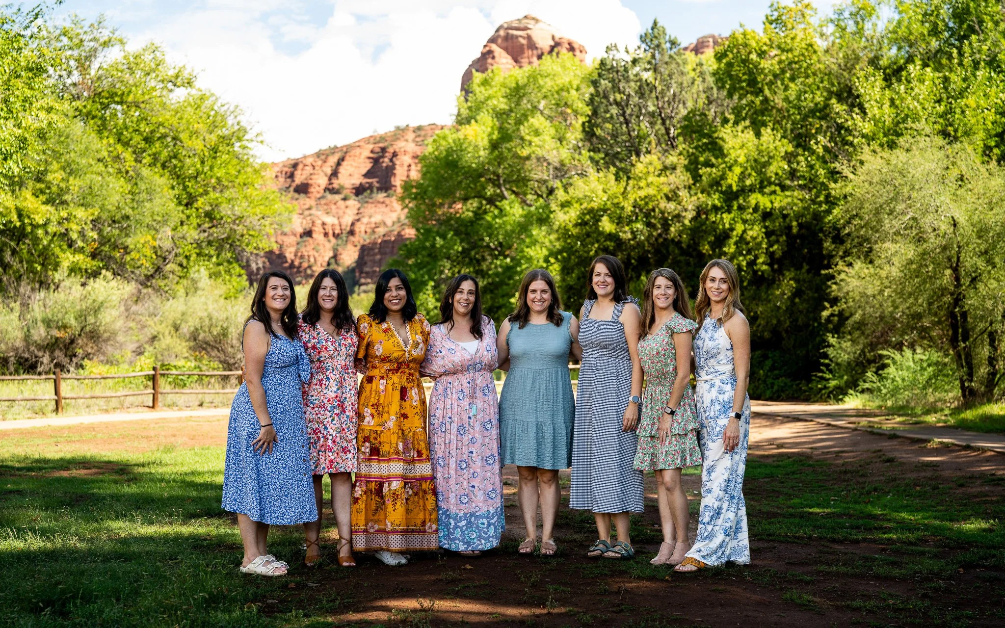 Group of eight women standing outdoors in a park with trees and red rock formations in the background, all wearing colorful summer dresses and smiling.