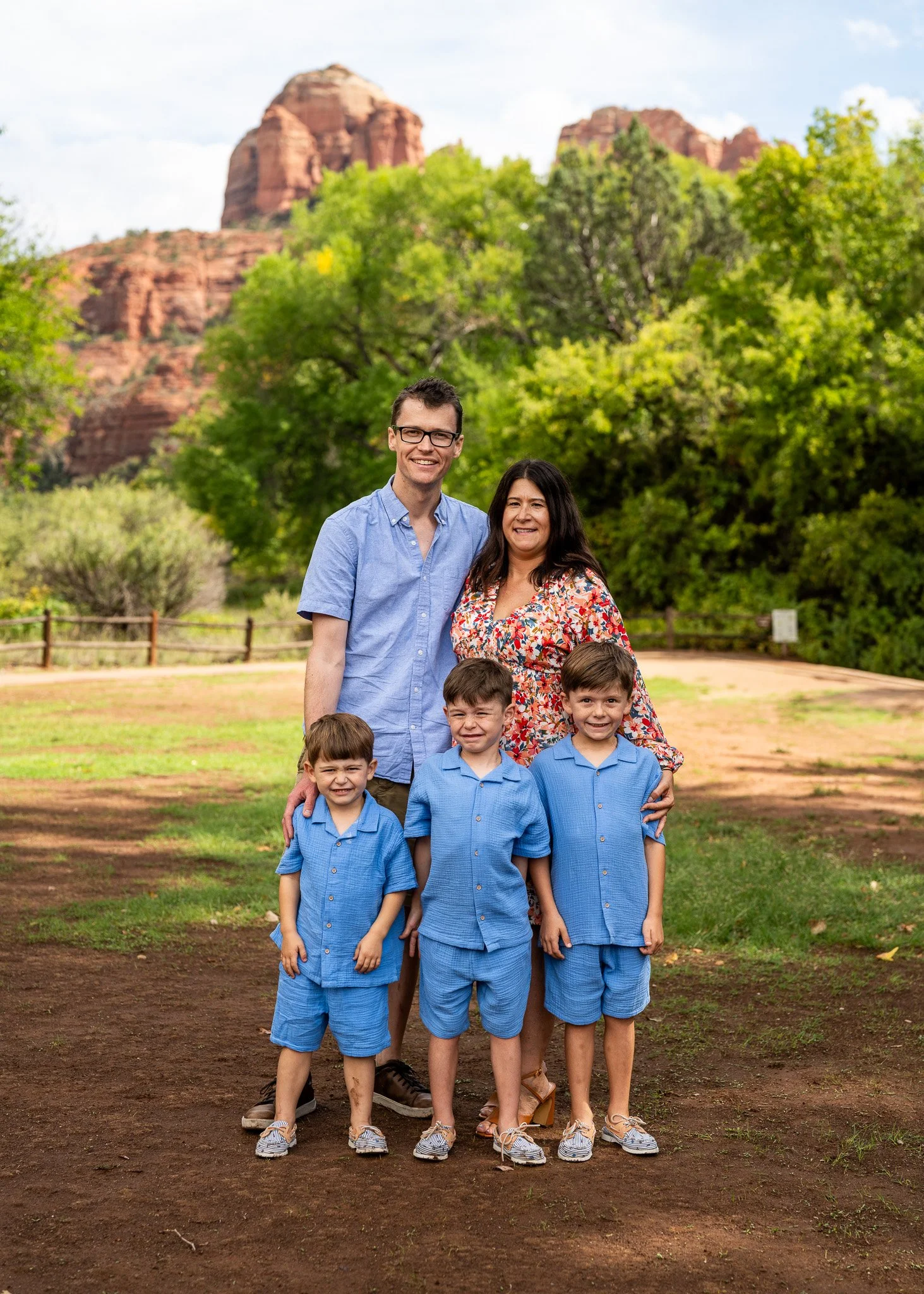 Family of six outdoors in a park, with red rock formations and green trees in the background, smiling for a photo.